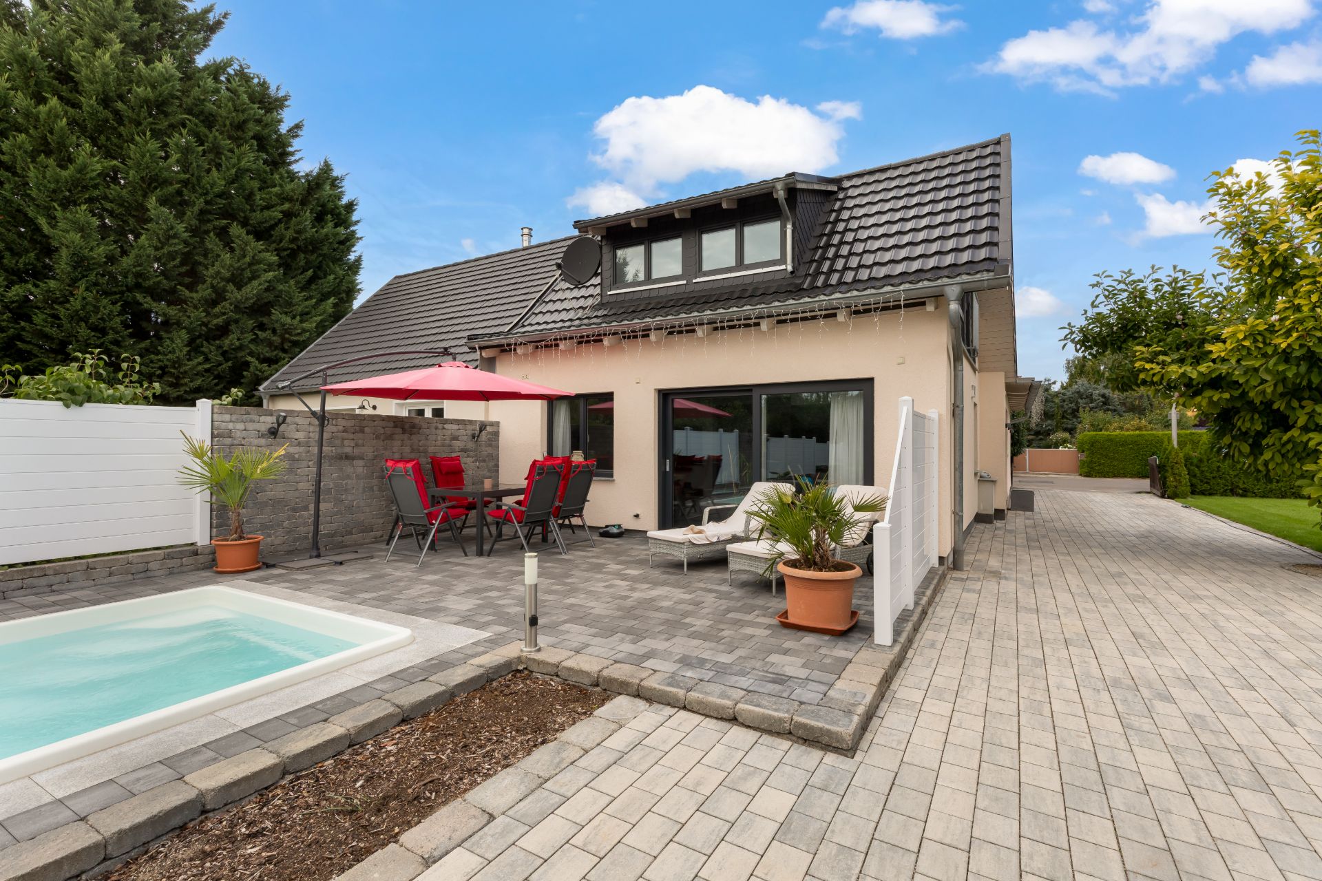 Gepflasterte Terrasse mit Sitzgruppe, Sonnenschirm und kleinem Pool an einem Einfamilienhaus.