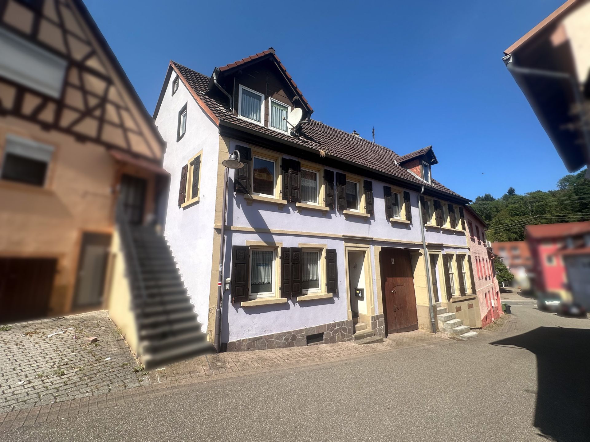 Zweistöckiges Wohnhaus mit hellvioletter Fassade und schwarzen Fensterläden an einer ruhigen Straße.