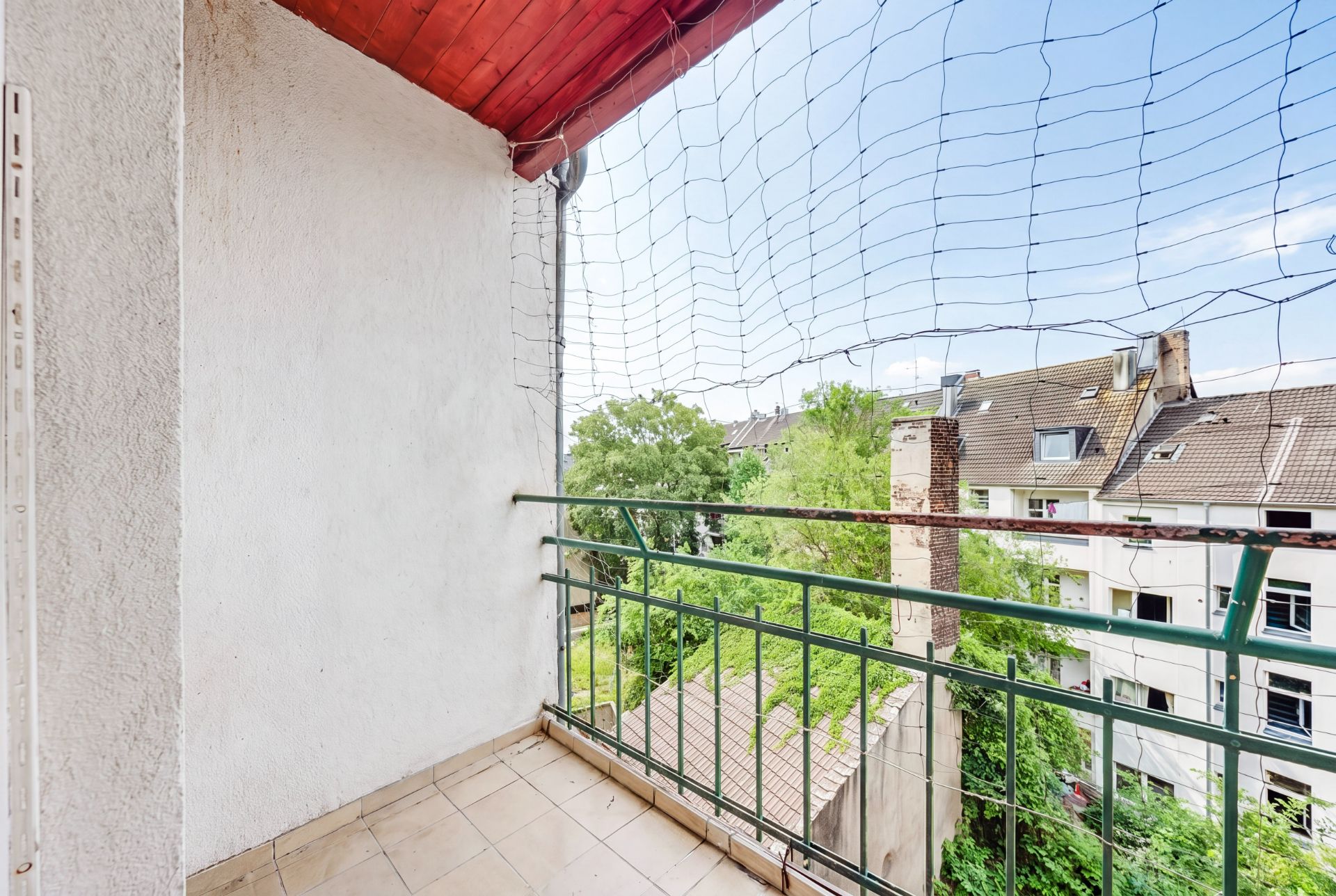 Kleiner Balkon mit Fliesenboden, grünem Geländer und Netz, Blick auf Bäume und Nachbarhäuser.