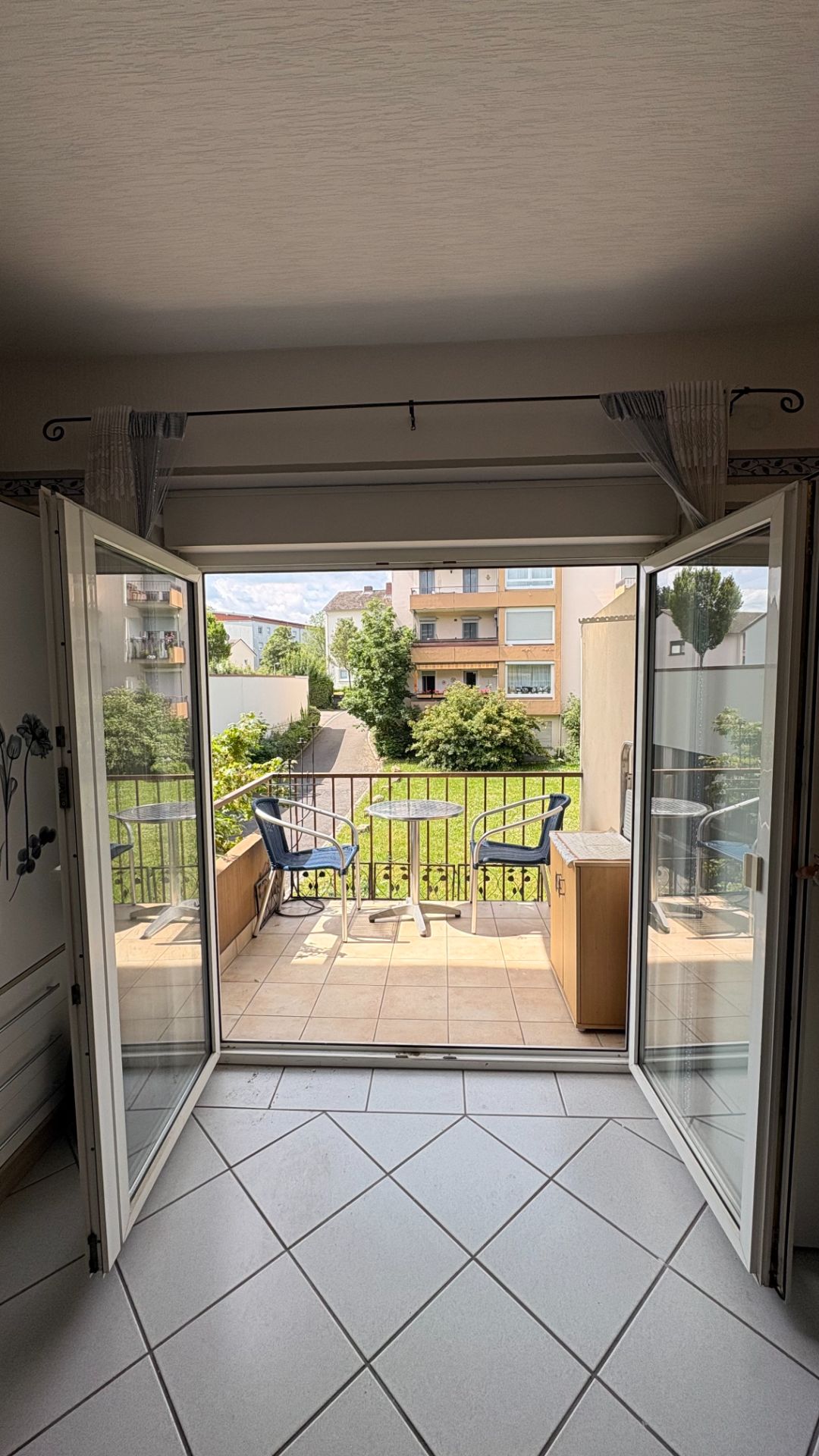 Offene Terrassentür mit Blick auf Balkon mit zwei Stühlen und Tisch, dahinter Garten und Wohnhäuser.
