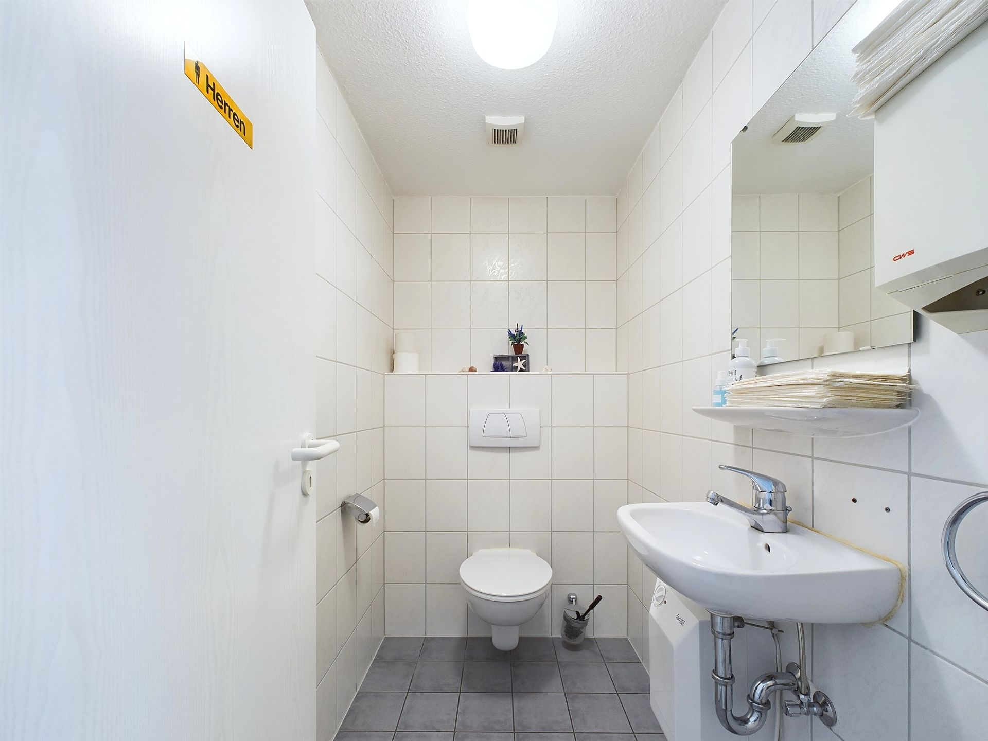 Kleines weiß gefliestes Badezimmer mit WC, Waschbecken, Spiegel und Ablage.