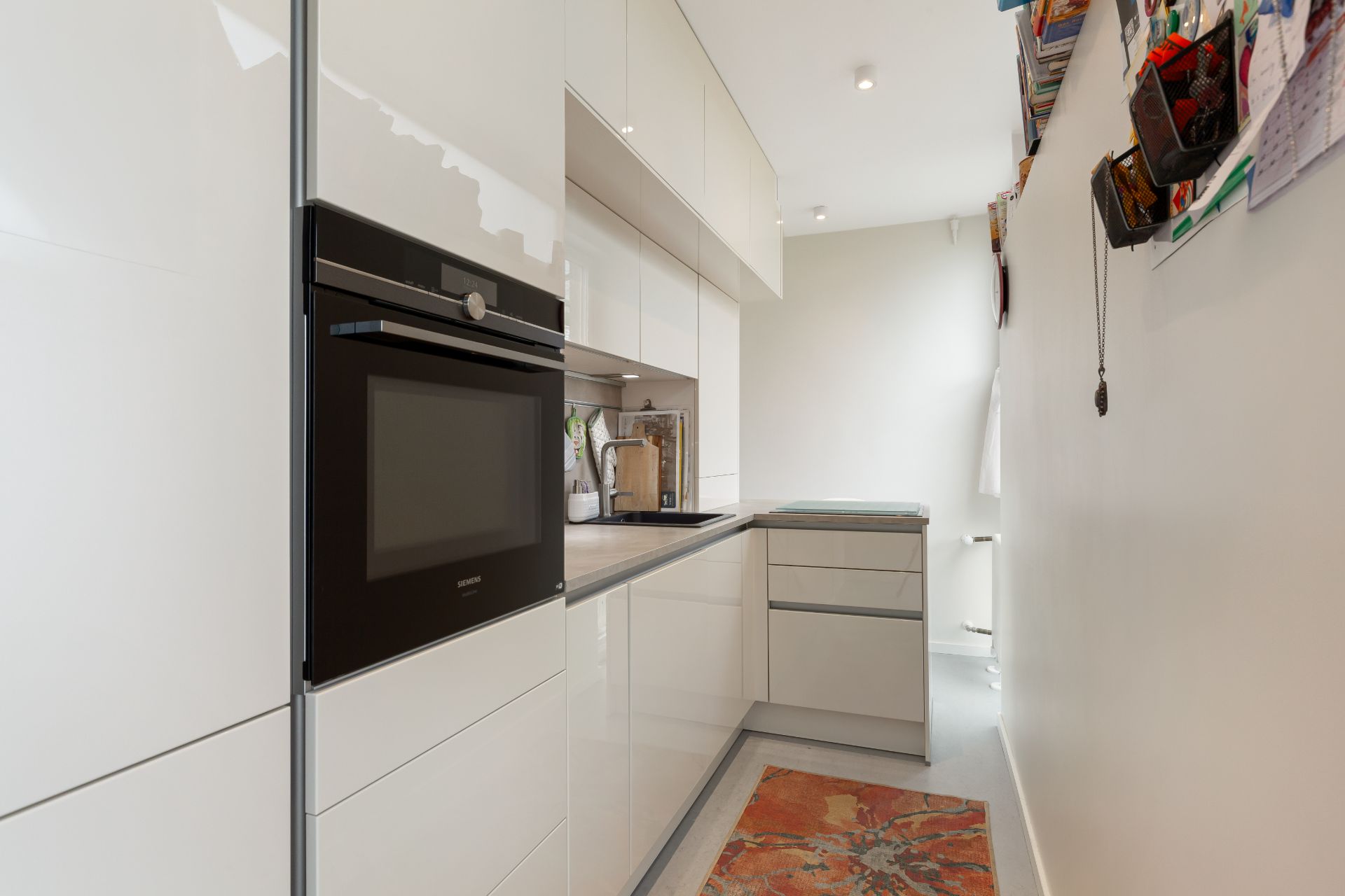 Moderne, schmale Küche mit weißen Hochglanzschränken, Einbauofen und kleinem Teppich am Boden.