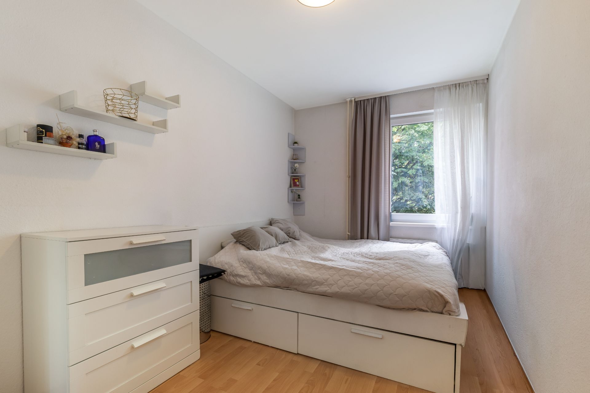 Helles Schlafzimmer mit Bett, Kommode und Fenster mit Vorhang.