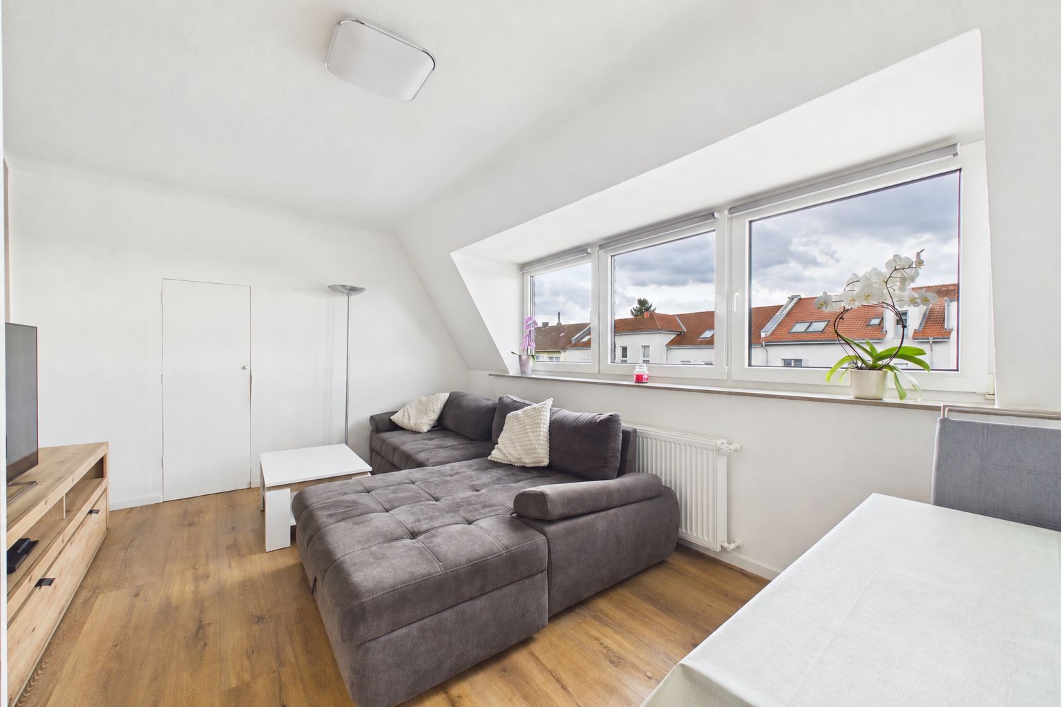 Helles Wohnzimmer mit grauem Ecksofa, Holzboden und großen Fenstern mit Blumen auf der Fensterbank.