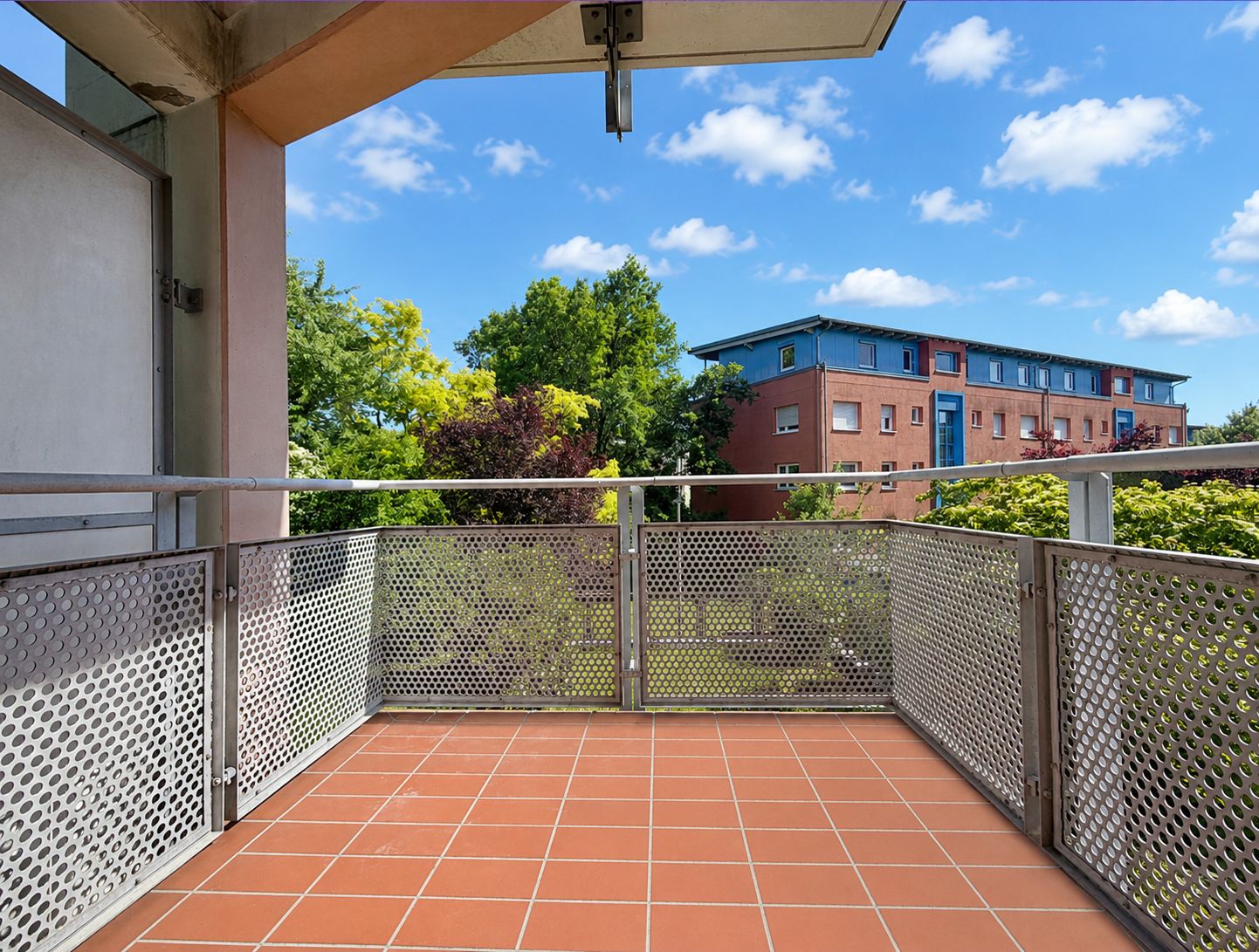 Balkon mit roten Fliesen und Metallgeländer, Blick auf Bäume und Wohngebäude.