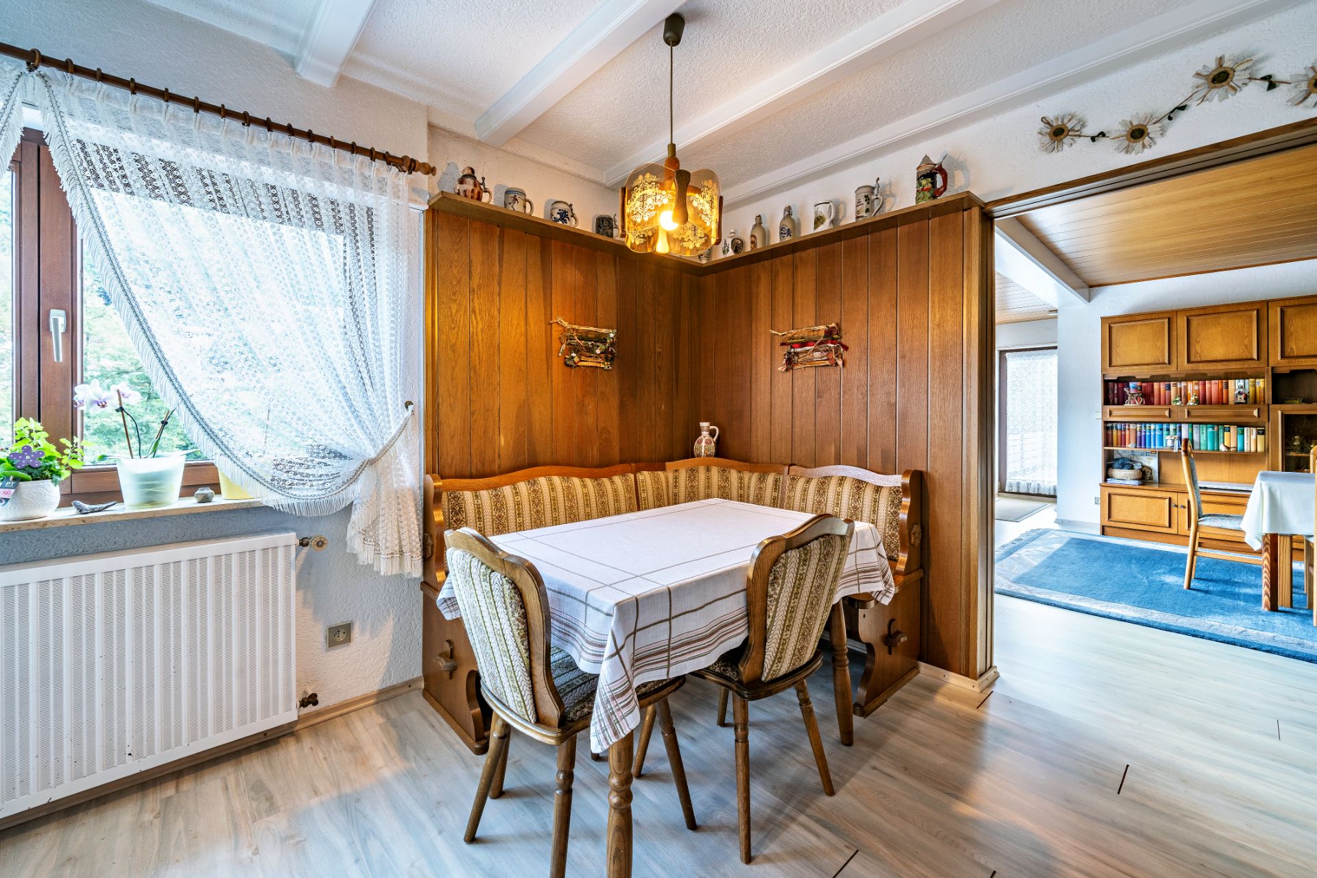 Gemütliche Sitzecke mit Holzwänden, Eckbank, Tisch und zwei Stühlen an einem Fenster mit Gardine.