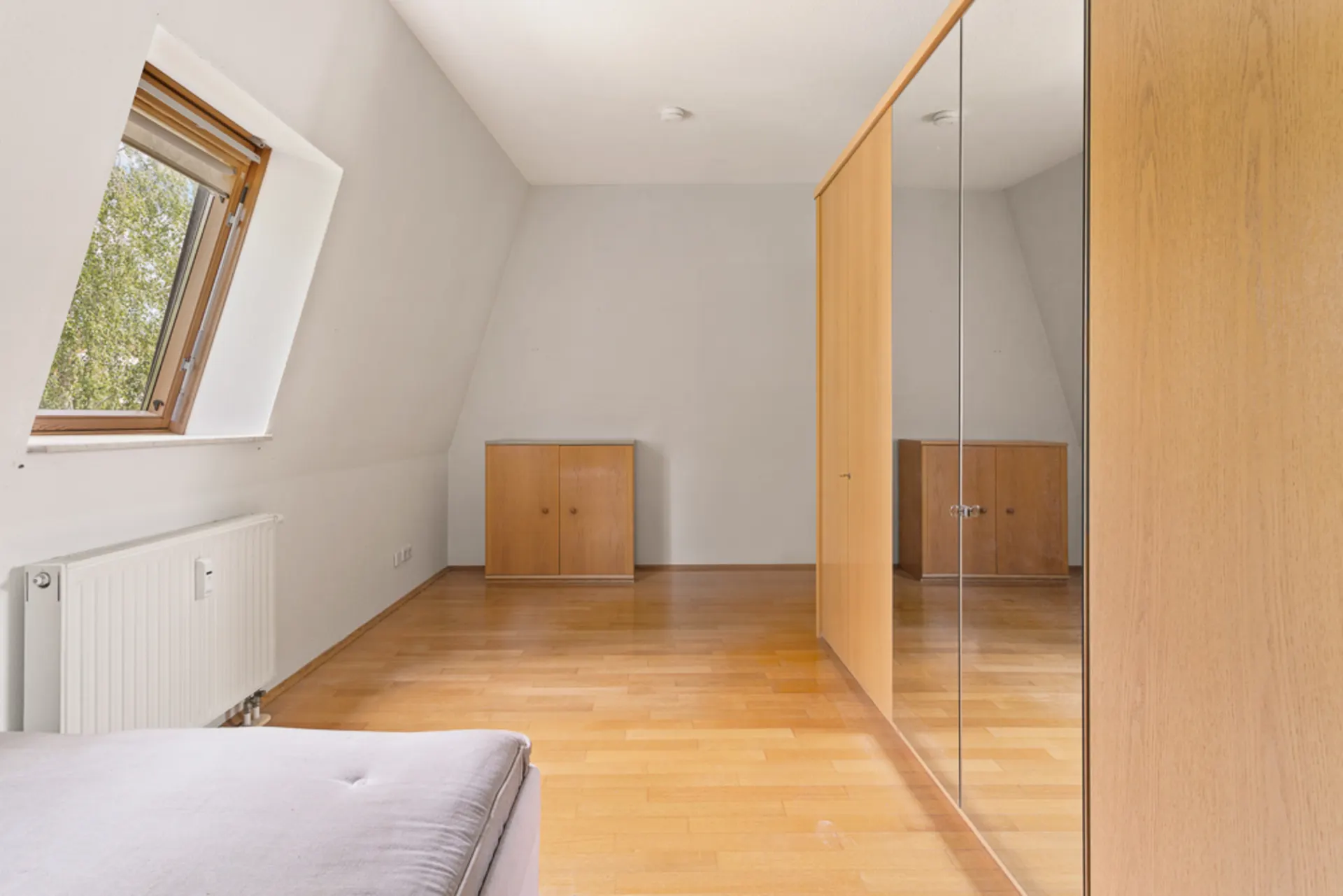 Helles Zimmer mit schräger Decke, Holzboden, Spiegelschrank und Fenster.