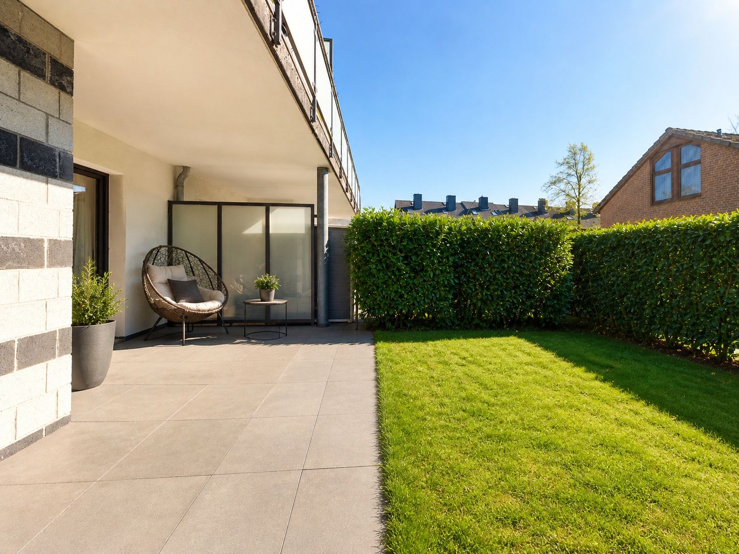 Moderne Terrasse mit Fliesenboden, Stuhl, Pflanzen und angrenzendem Rasen, umgeben von Hecken.