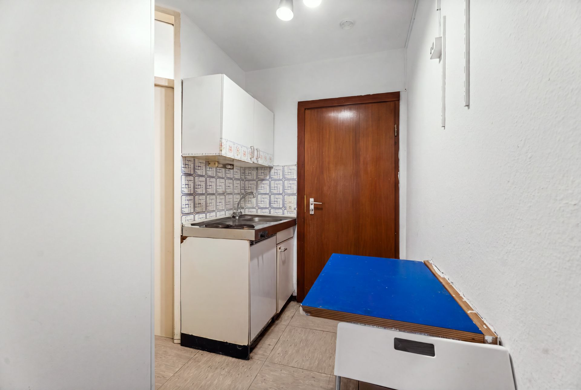 Kleine Küche mit Spüle, Hängeschrank, Holztür und blauem Tisch an der Wand.