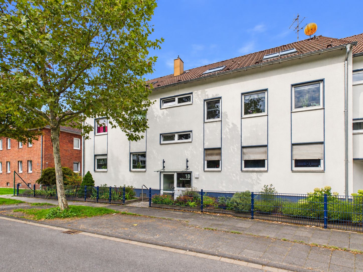Wohnhaus mit weißer Fassade, mehreren Fenstern und kleinem Vorgarten, davor ein Gehweg und ein Baum.