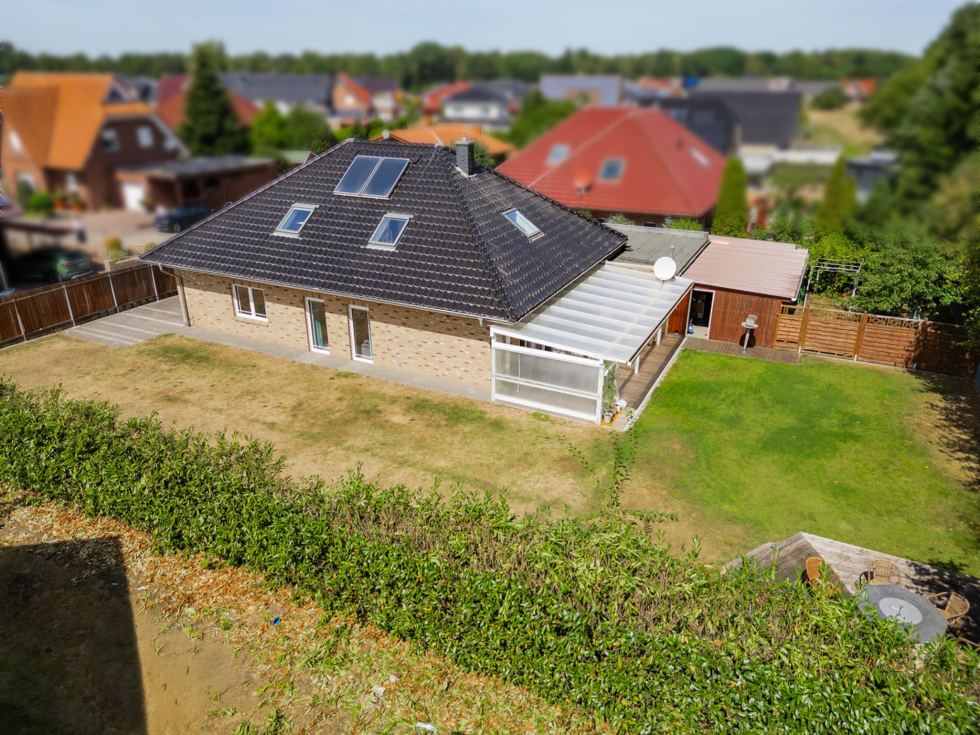 Einfamilienhaus mit dunklem Dach, Wintergarten und großem Garten, umgeben von Hecke und Zaun.