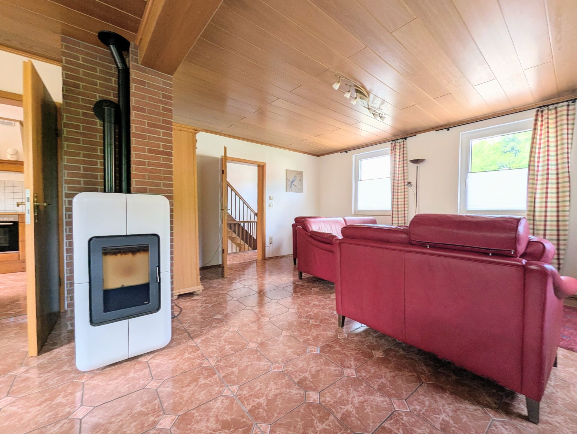 Wohnzimmer mit roten Sofas, Kaminofen, Fliesenboden und zwei Fenstern mit Vorhängen.