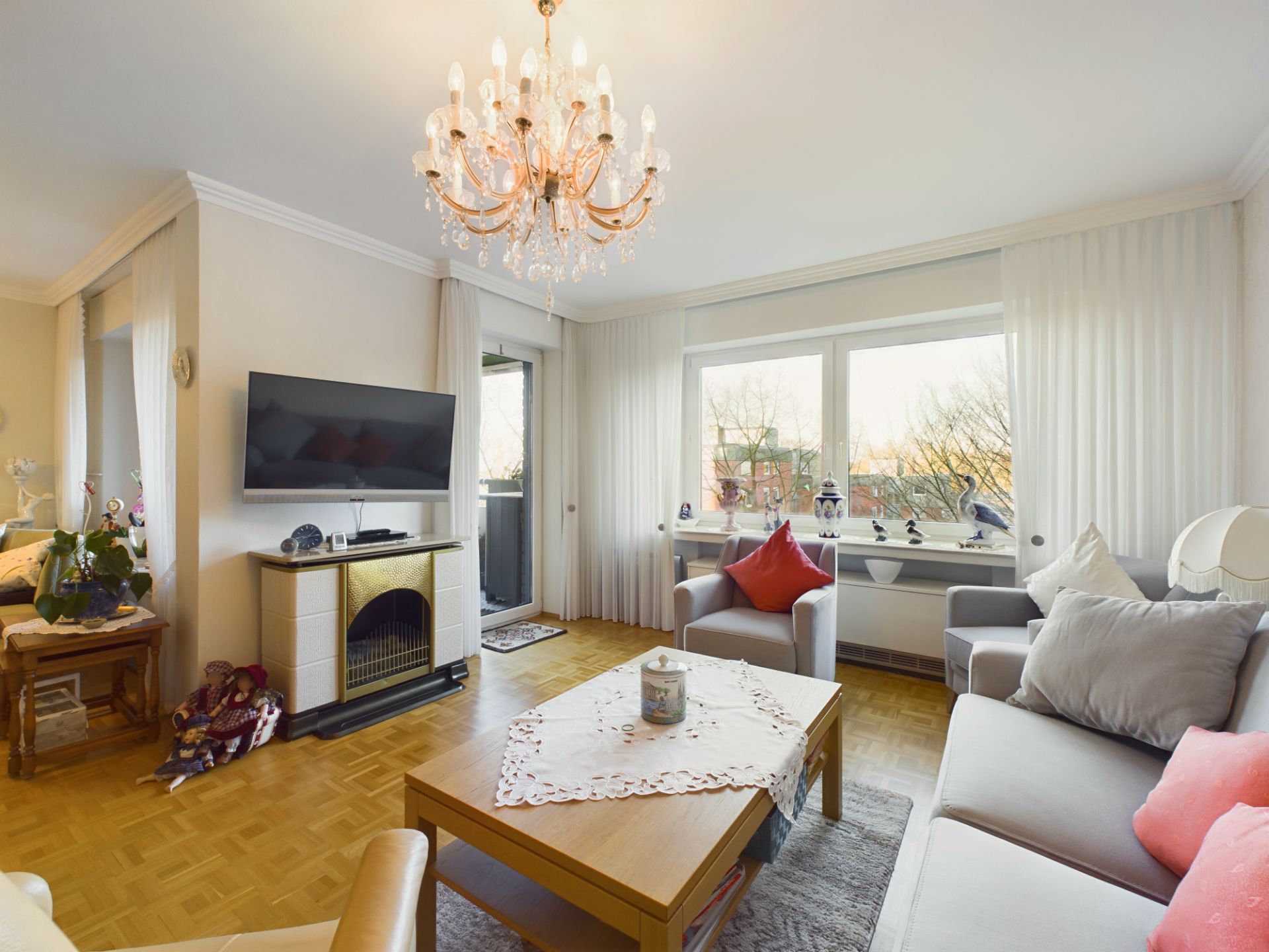 Helles Wohnzimmer mit Sofa, Sesseln, Couchtisch, großem Fenster und Kronleuchter.