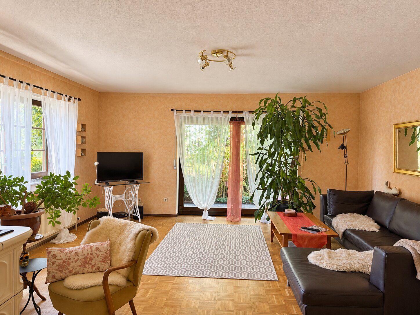 Helles Wohnzimmer mit Sofa, Sessel, Teppich, mehreren Pflanzen und großem Fenster mit Vorhängen.