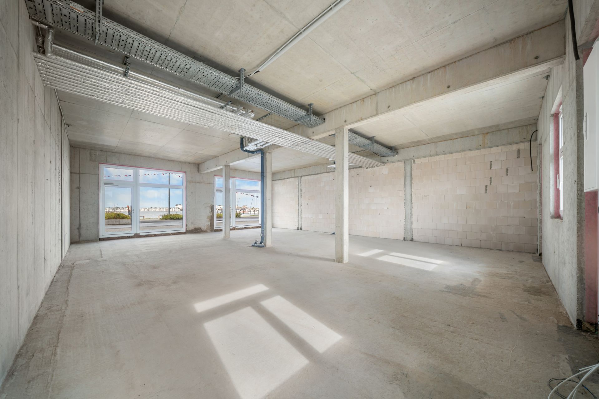 Leerer Raum mit Sichtbetonwänden, großen Fenstern und sichtbaren Leitungen an der Decke.