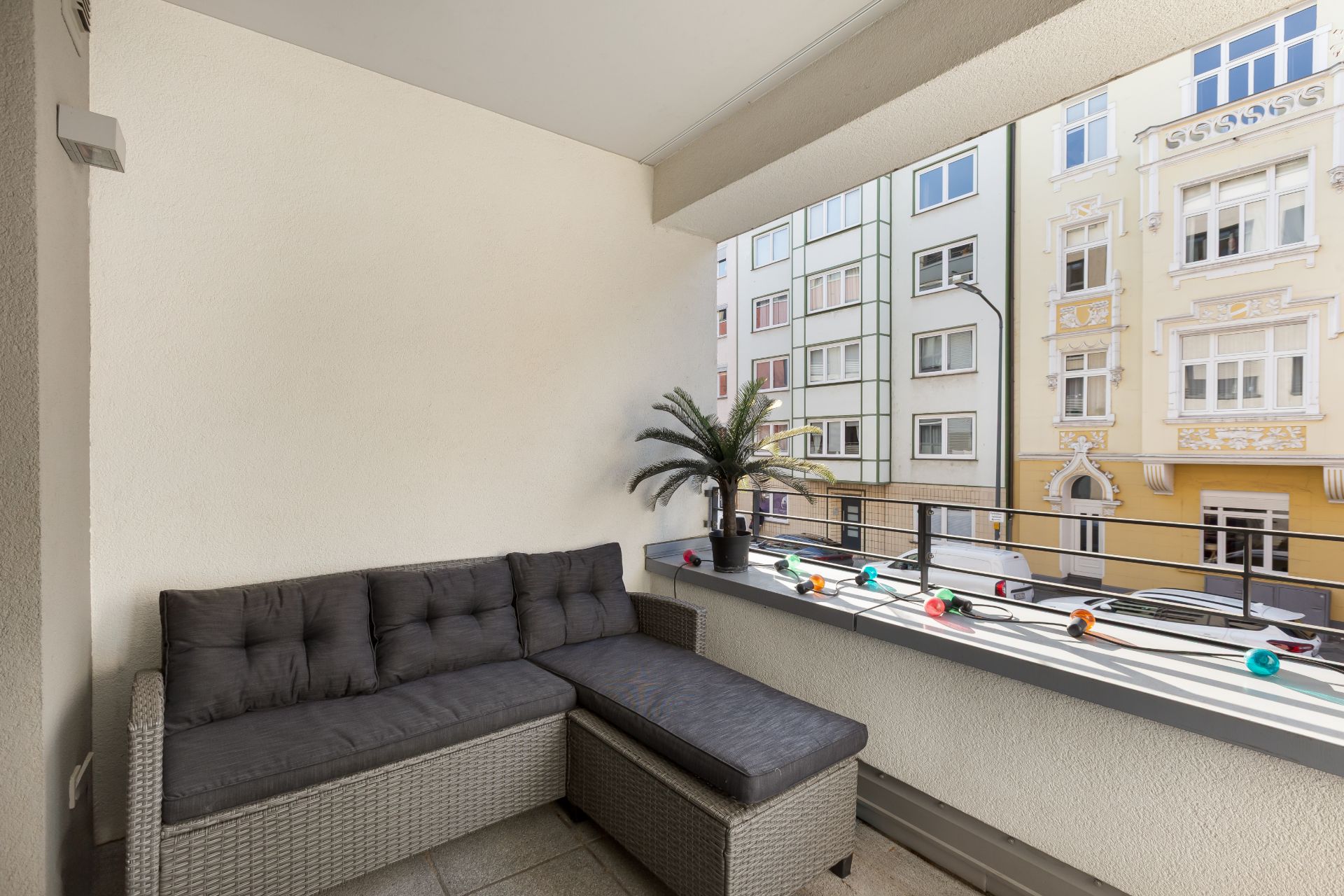 Überdachter Balkon mit grauem Sofa, kleiner Palme und bunten Lichterketten, Blick auf Nachbarhäuser.