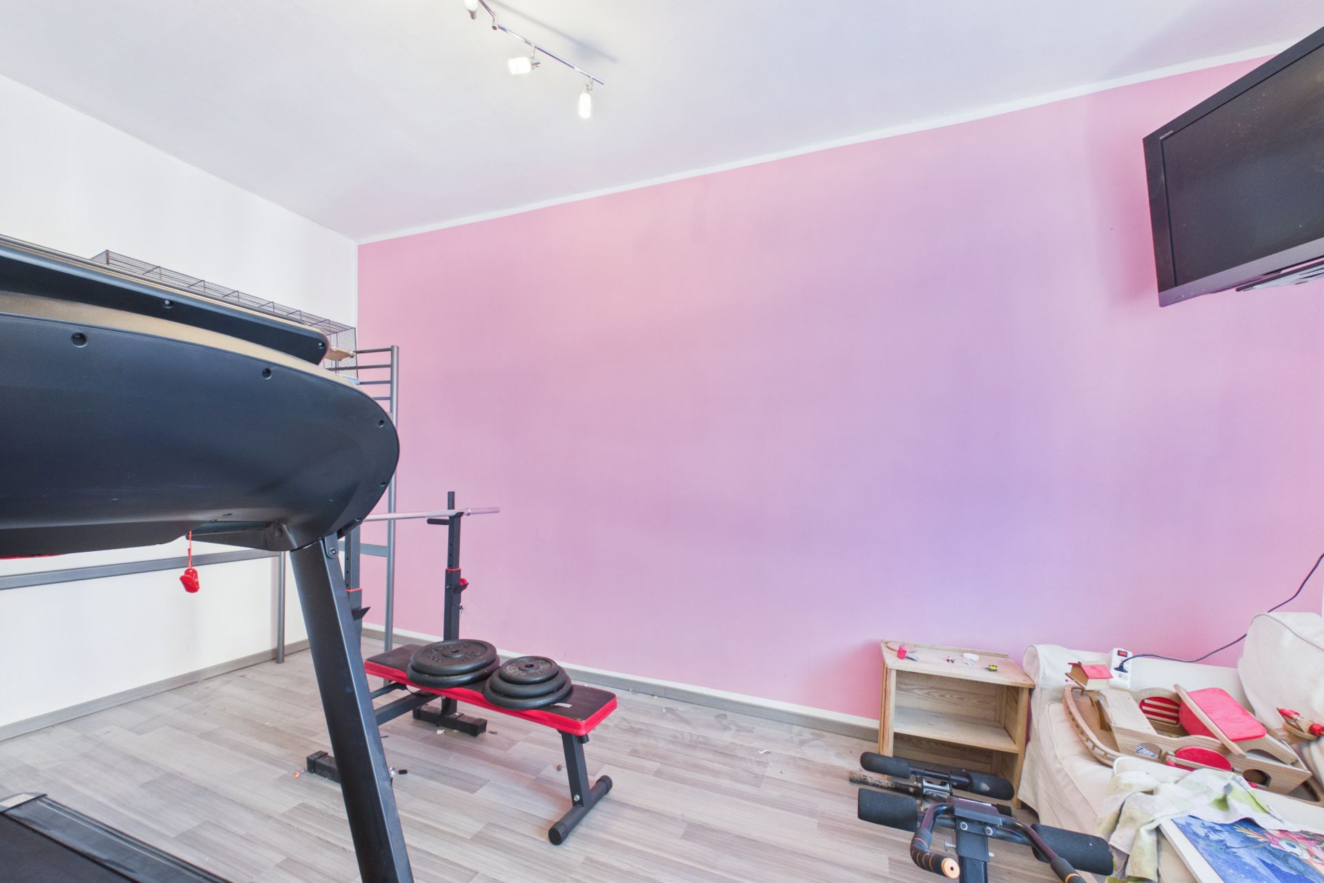 Fitnessraum mit Laufband, Hantelbank, Gewichten und Fernseher an der Wand.