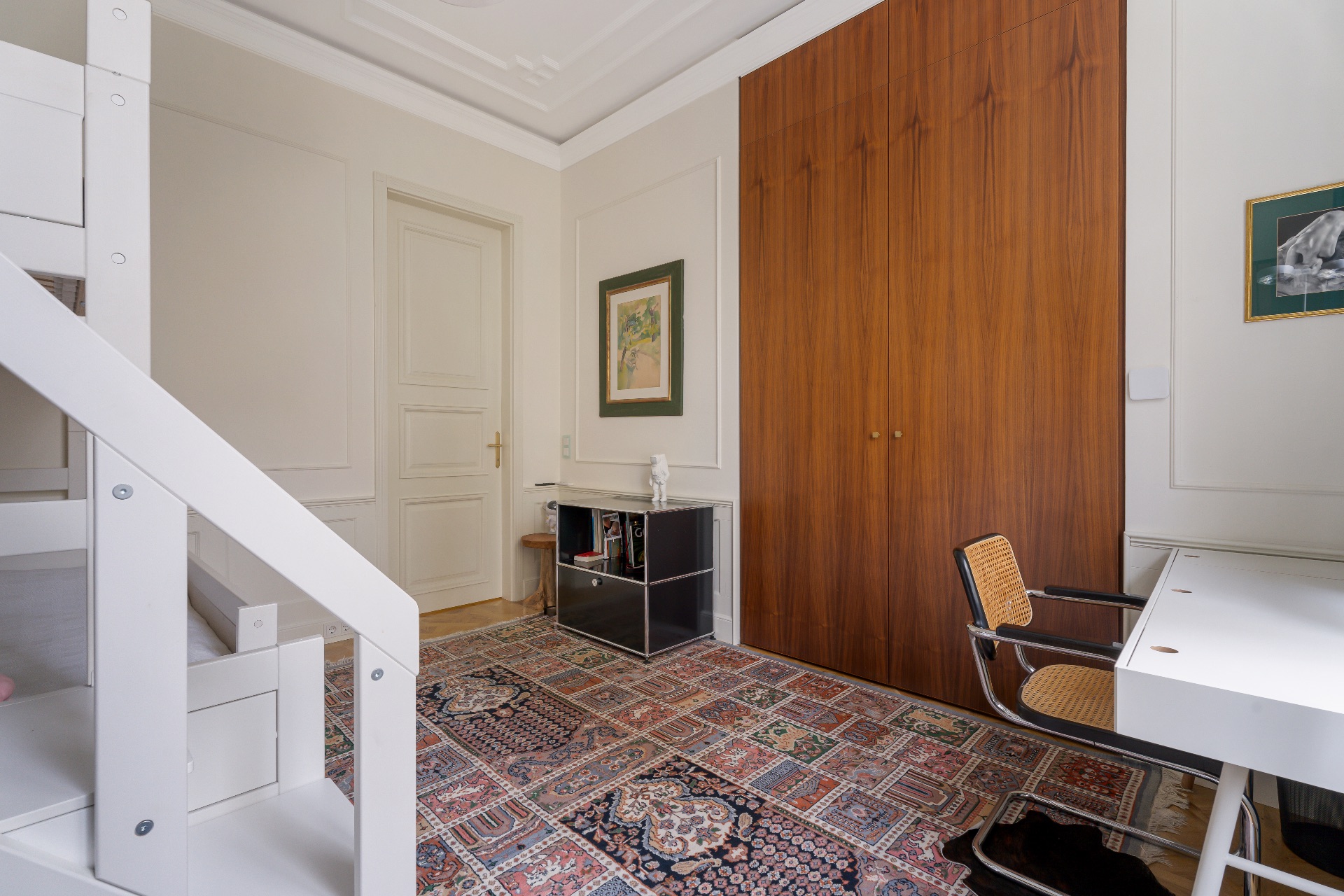 Helles Zimmer mit Hochbett, Schreibtisch, Stuhl, Teppich und großem Holzschrank.