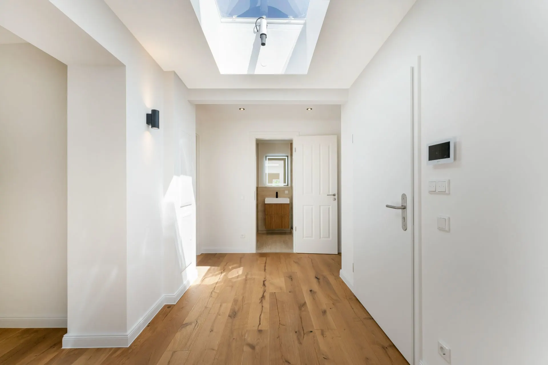 Heller Flur mit Holzfußboden, Dachfenster und Blick auf ein Badezimmer mit Waschbecken.