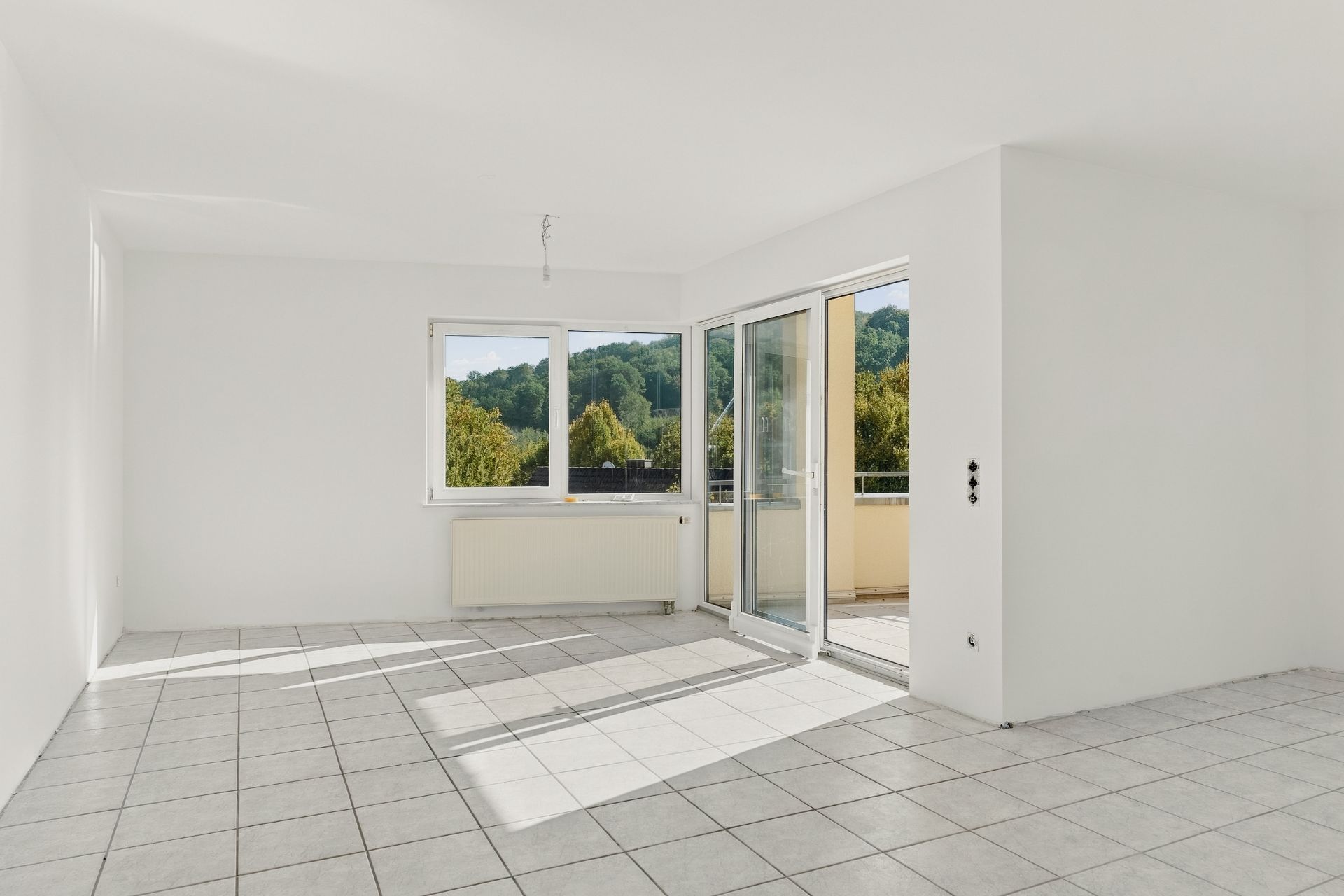 Helles, leeres Zimmer mit Fliesenboden, großen Fenstern und Zugang zu einem Balkon.