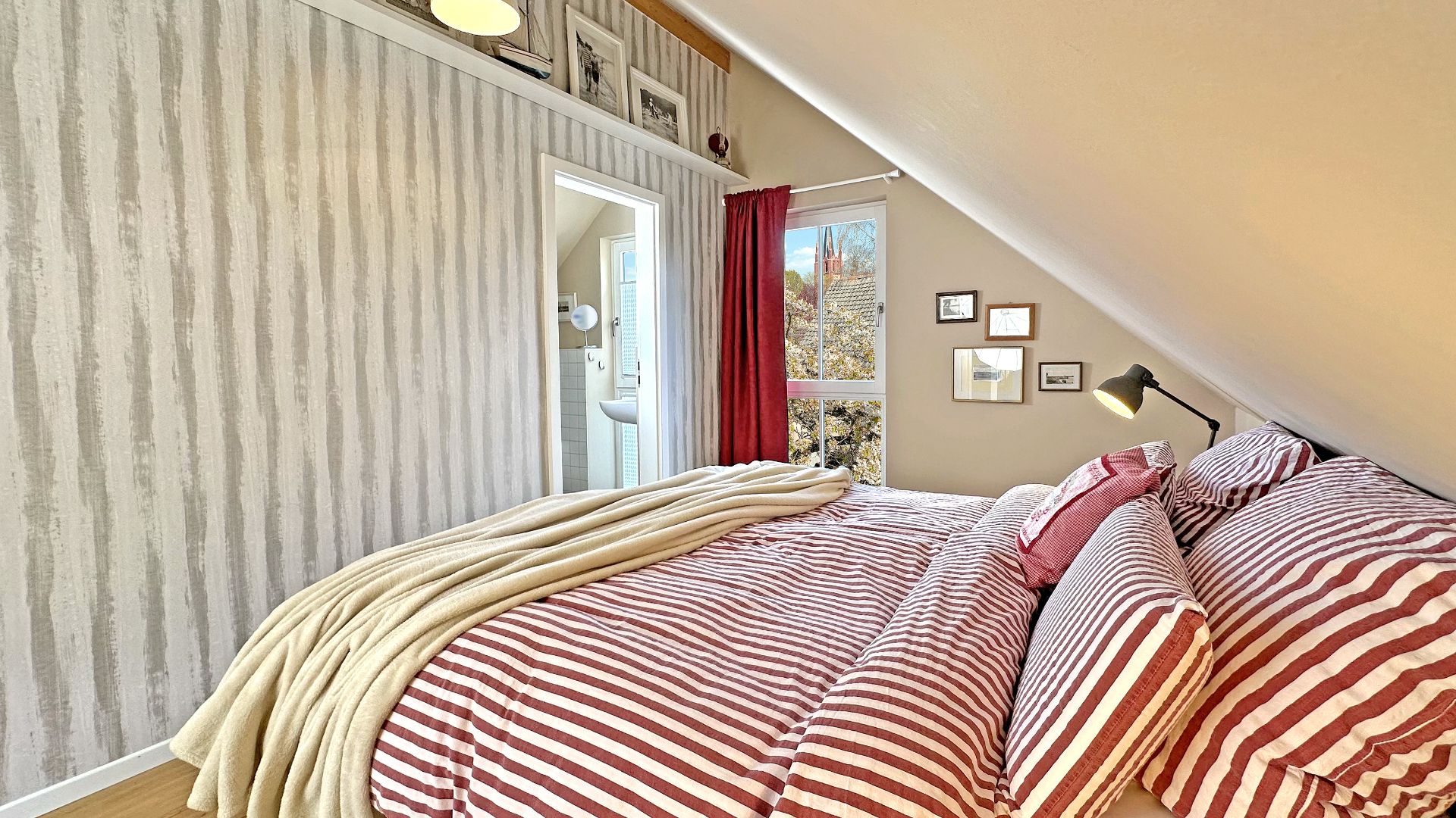 Schlafzimmer mit Dachschräge, gestreiftem Bett, Wandlampen und Zugang zu einem kleinen Bad.