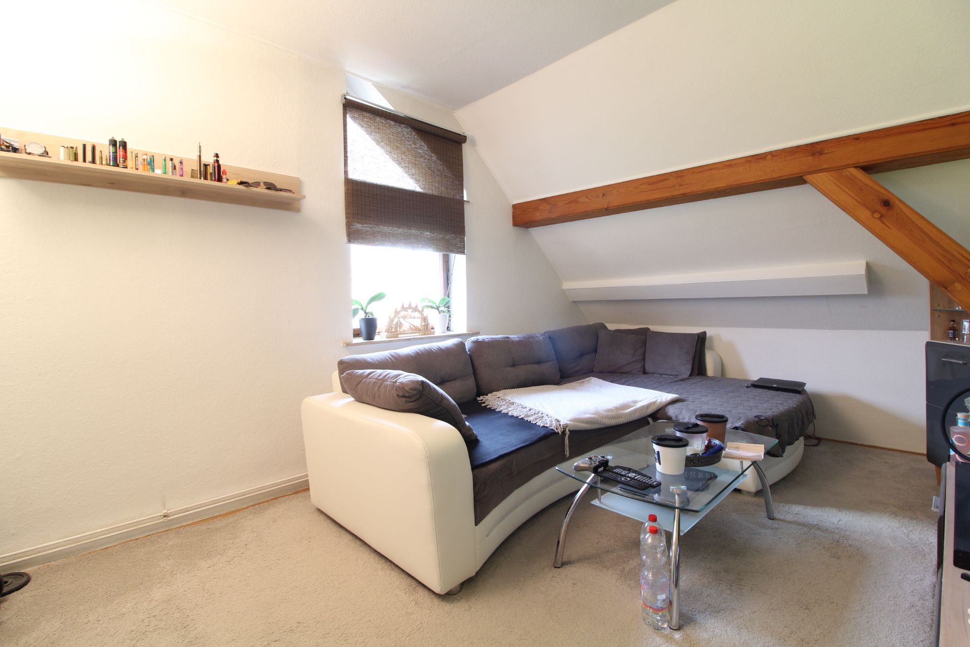 Wohnzimmer mit Ecksofa, Glastisch, Regal an der Wand und Fenster mit Rollo.