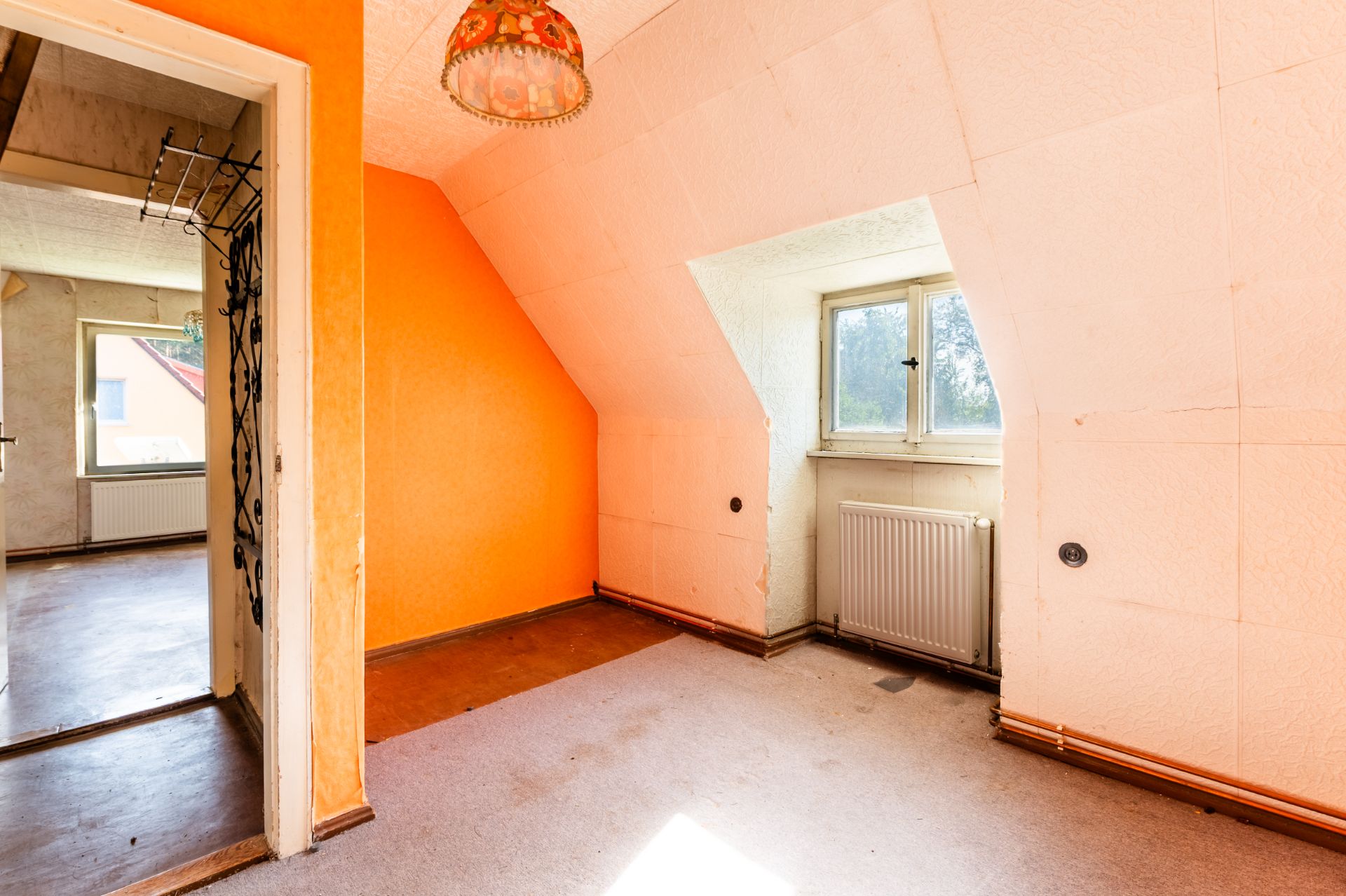 Helles Zimmer mit orangefarbener Wand, Teppichboden, Fenster und Heizkörper.