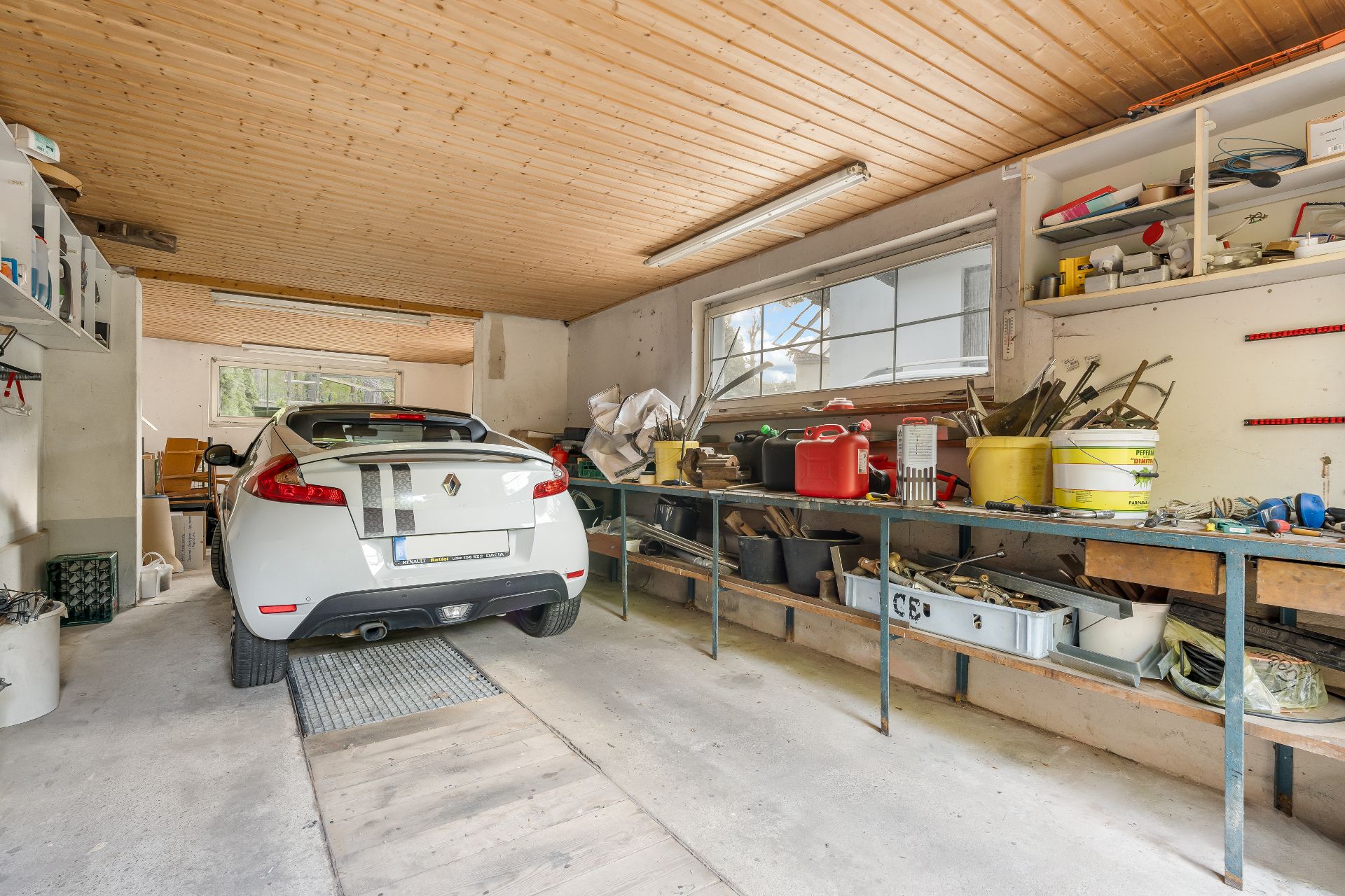 Garage mit geparktem Auto, Werkbänken, Werkzeugen und Regalen an den Wänden.