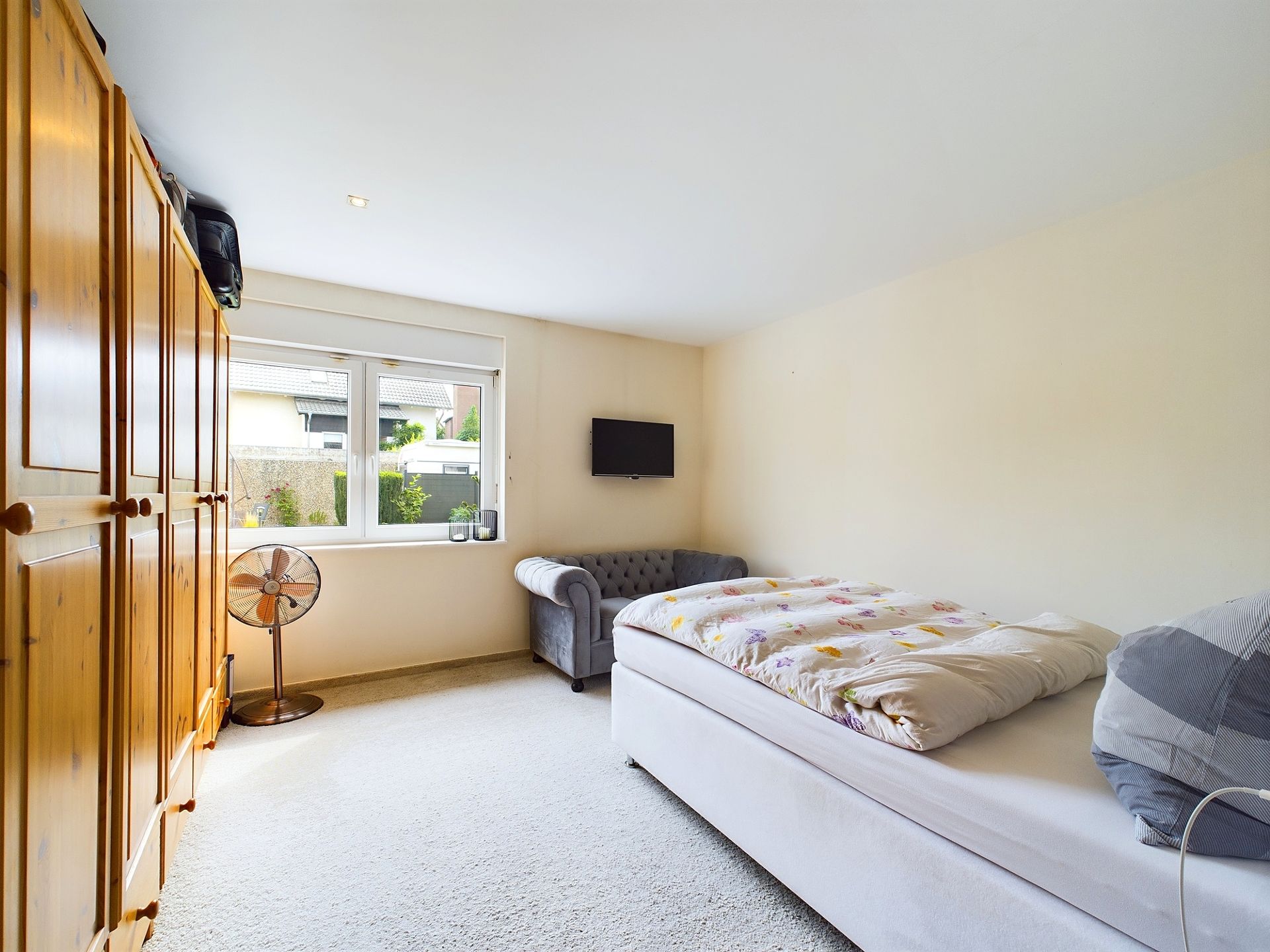 Helles Schlafzimmer mit Bett, grauem Sofa, Holzschrank und Fenster.