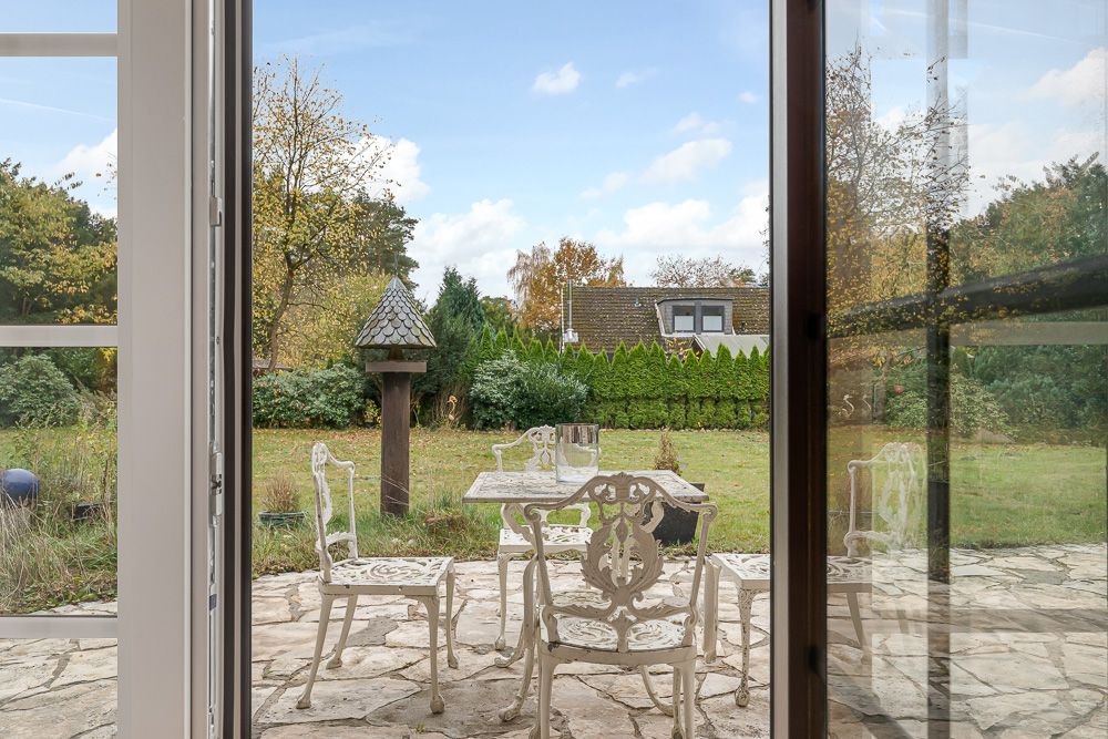 Blick durch eine Glastür auf eine Terrasse mit weißem Metalltisch und Stühlen im Garten.