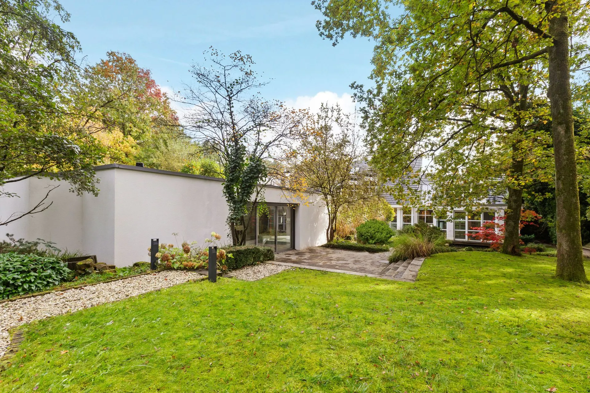 Garten mit Rasenfläche, Bäumen und einem weißen, modernen Flachbauhaus im Hintergrund.