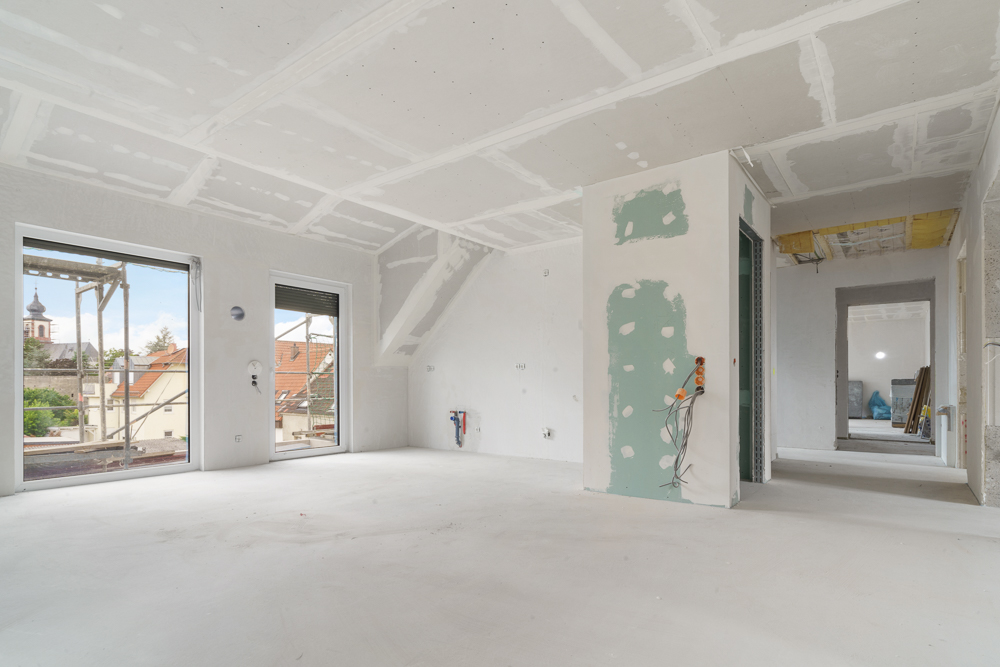 Heller Raum im Rohbau mit zwei großen Fenstern und sichtbaren Kabeln an der Wand.