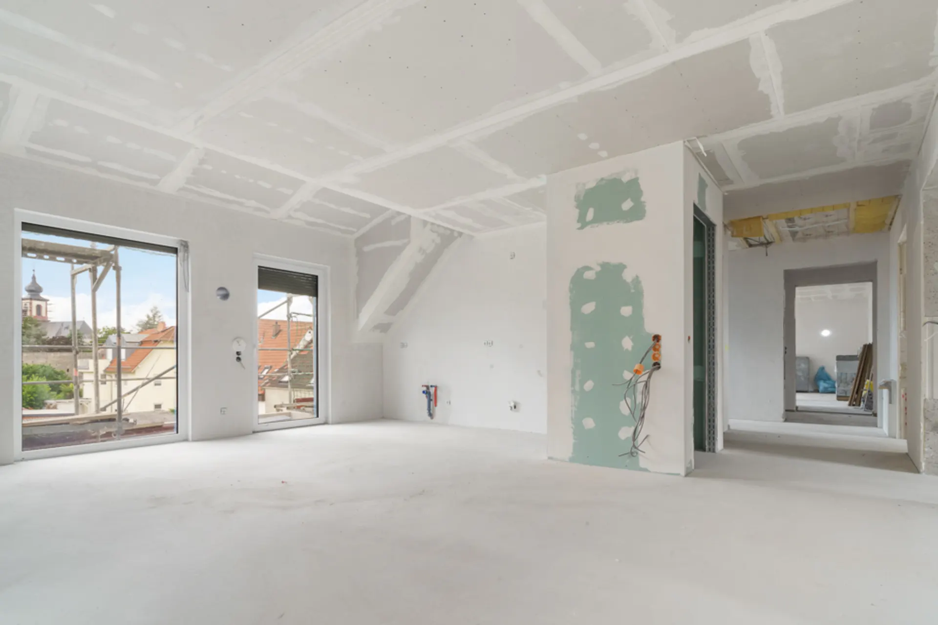 Heller Raum im Rohbau mit zwei großen Fenstern und sichtbaren Kabeln an der Wand.