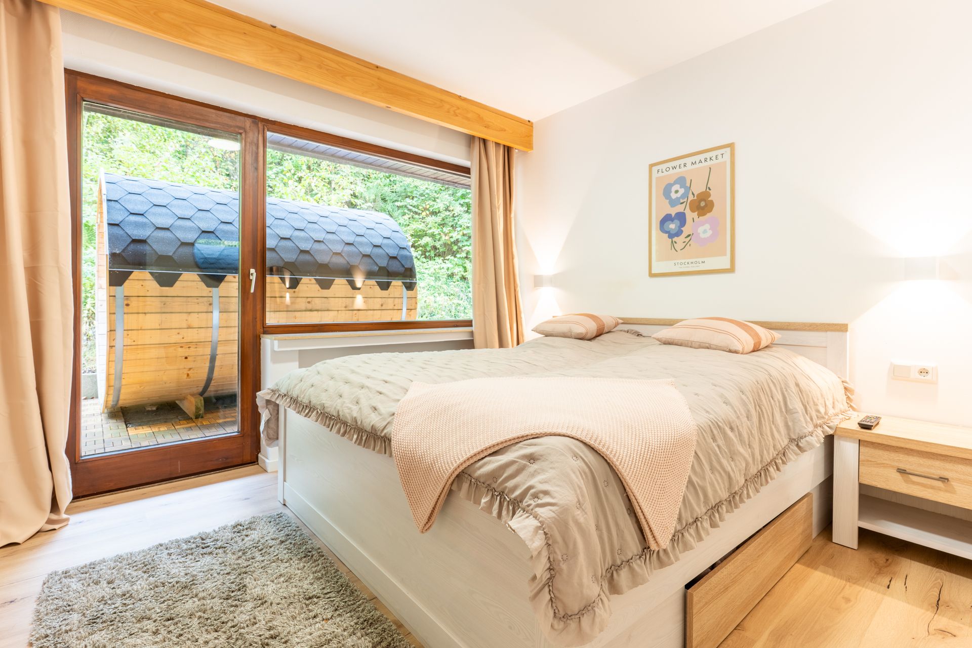 Helles Schlafzimmer mit großem Bett, Nachttisch, Wandbild und Blick auf Terrasse mit Holzsauna.