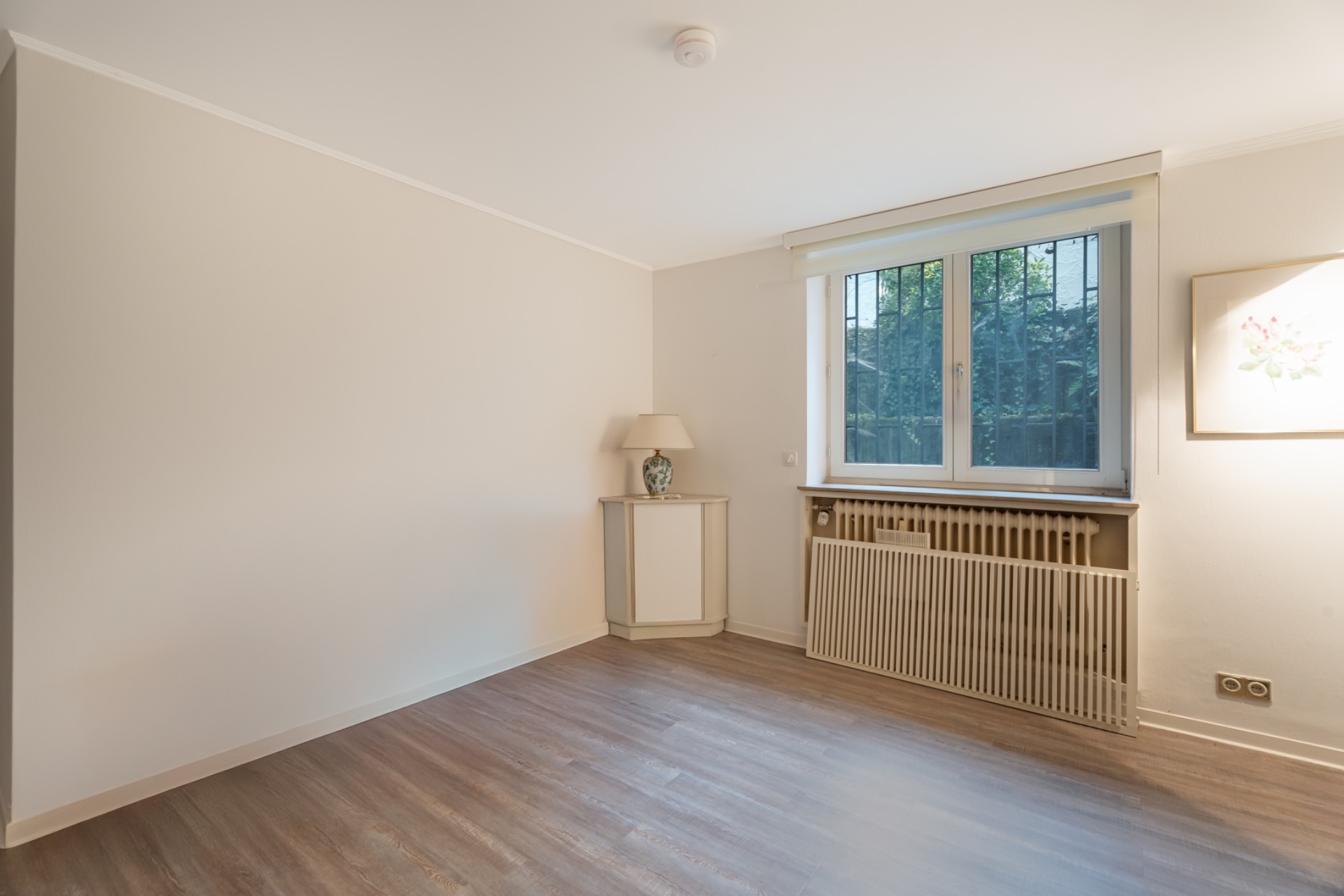 Leerer Raum mit Holzboden, Fenster, Heizung und kleiner Lampe auf einer Kommode.