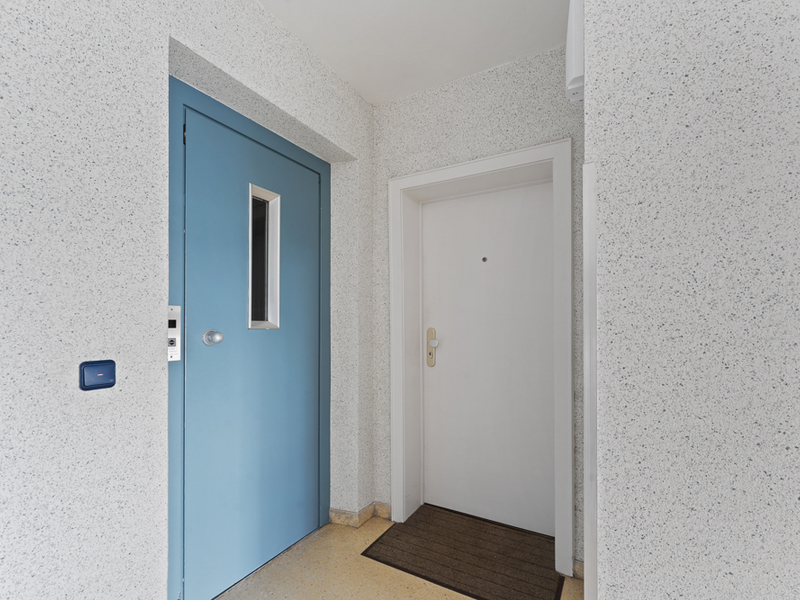 Flur mit blauem Aufzug und weißer Wohnungstür, davor liegt eine braune Fußmatte.