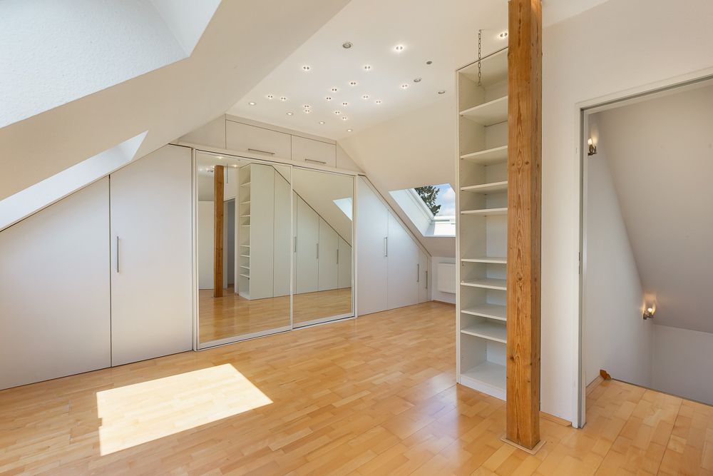 Heller Dachzimmer mit Holzfußboden, Einbauschränken, Spiegelwand und Regal neben Holzbalken.