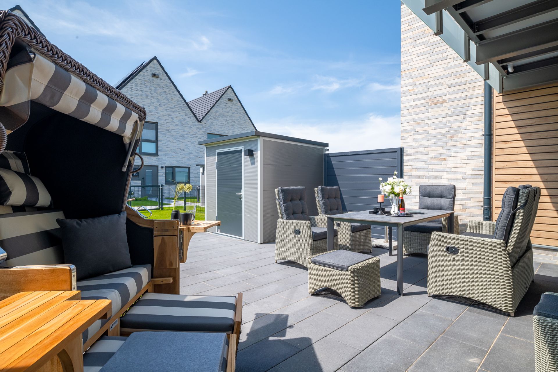 Moderne Terrasse mit Korbstühlen, Tisch und Strandkorb, im Hintergrund ein Gartenhaus.