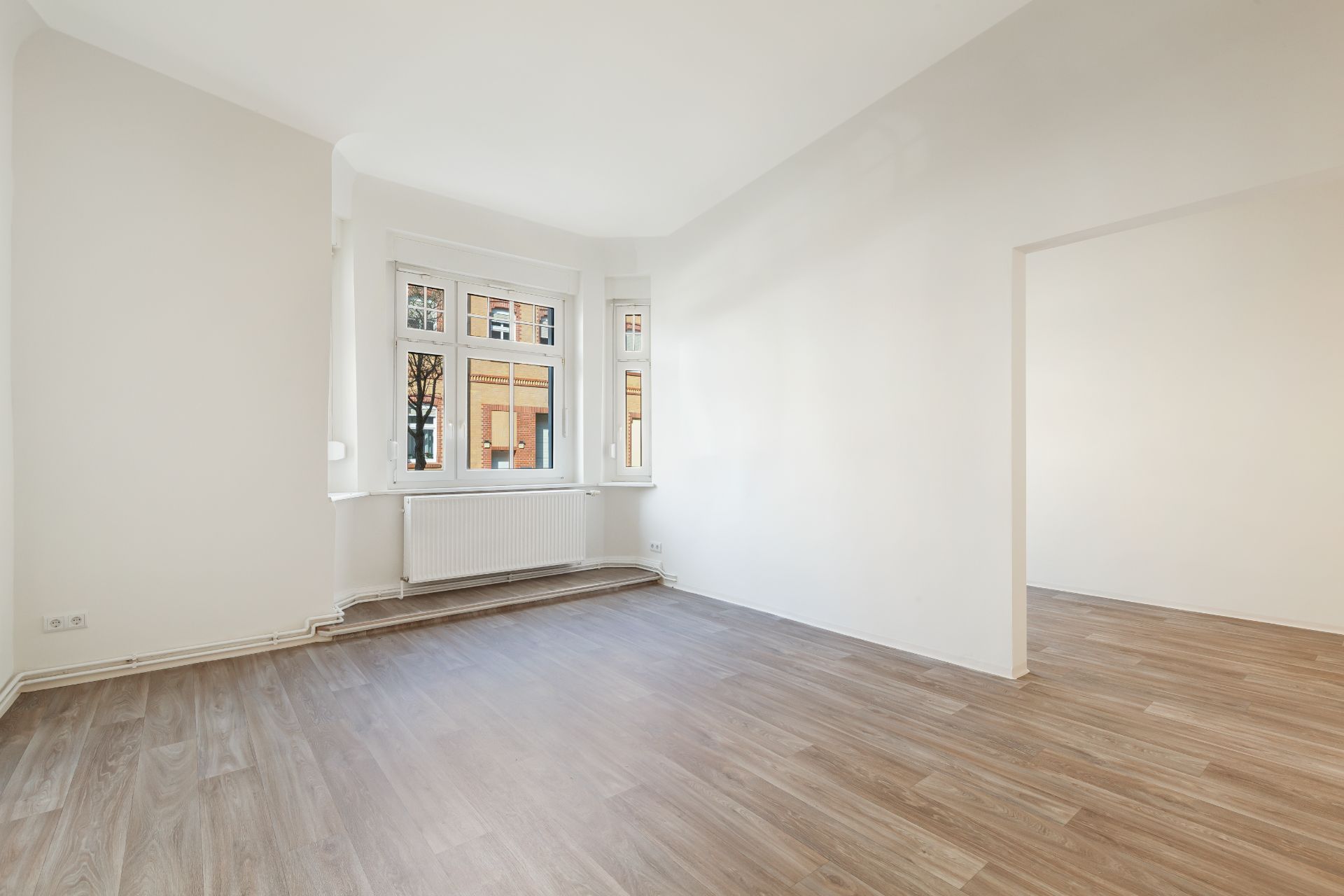 Leerer, heller Raum mit Holzfußboden und drei Fenstern, Blick auf Nachbargebäude.