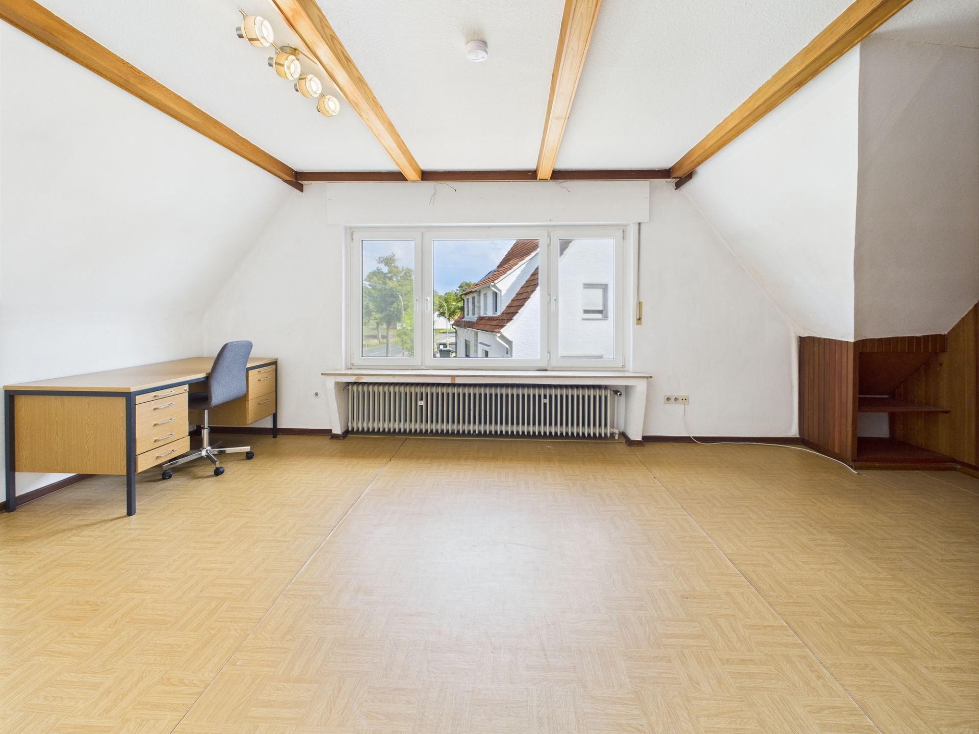 Heller Raum mit Schreibtisch, Stuhl, großem Fenster und sichtbaren Holzbalken an der Decke.
