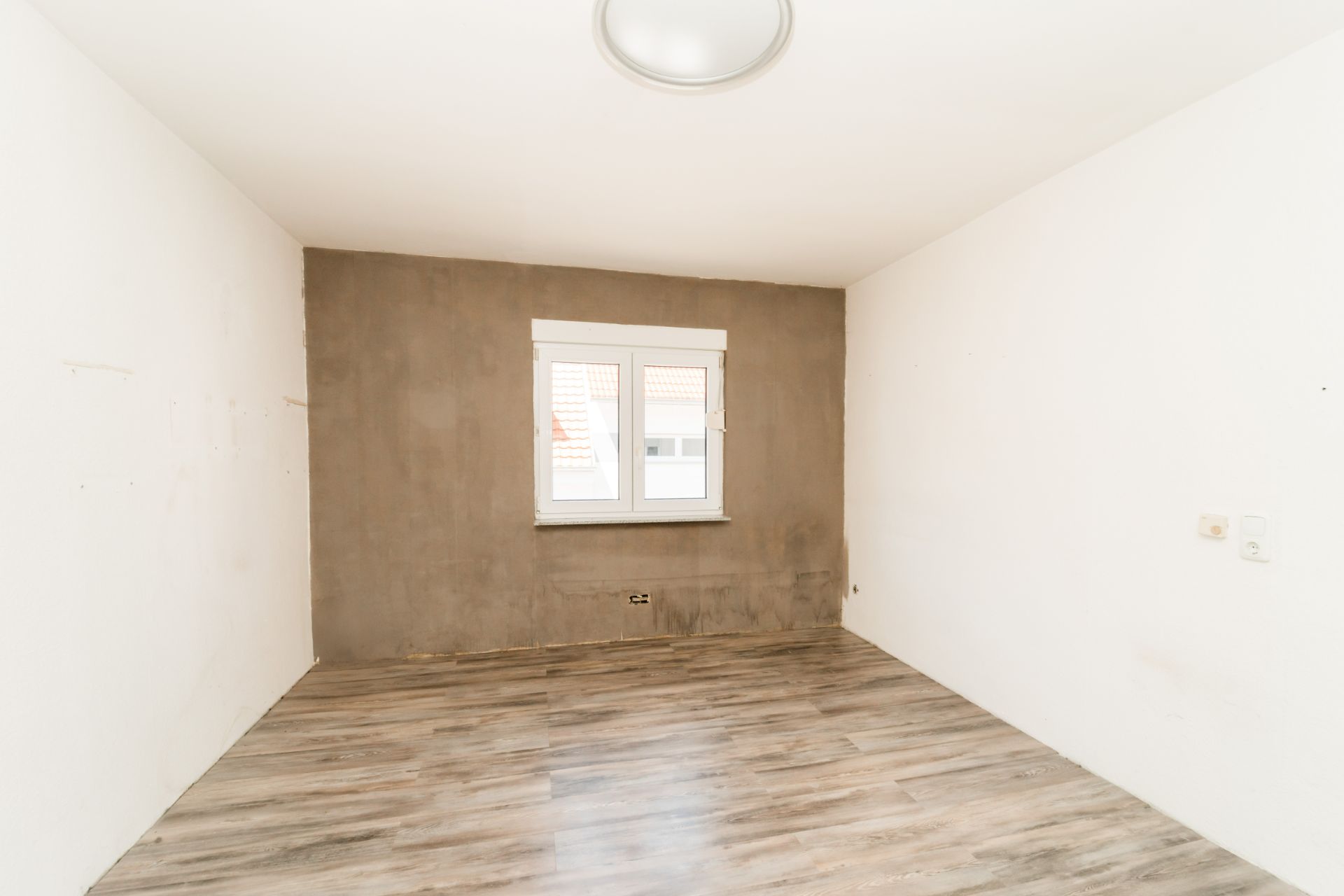 Leerer Raum mit Holzboden, weiß gestrichenen Wänden und einem Fenster an der Stirnseite.