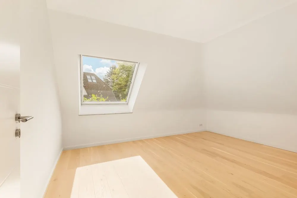 Leerer, heller Raum mit Holzfußboden und schrägem Fenster in einer weißen Wand.