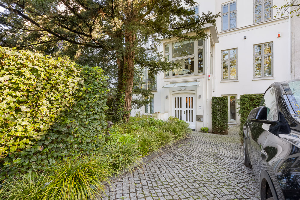 Eingang eines weißen Wohnhauses mit Kopfsteinpflasterweg, Auto und viel Grün vorne.