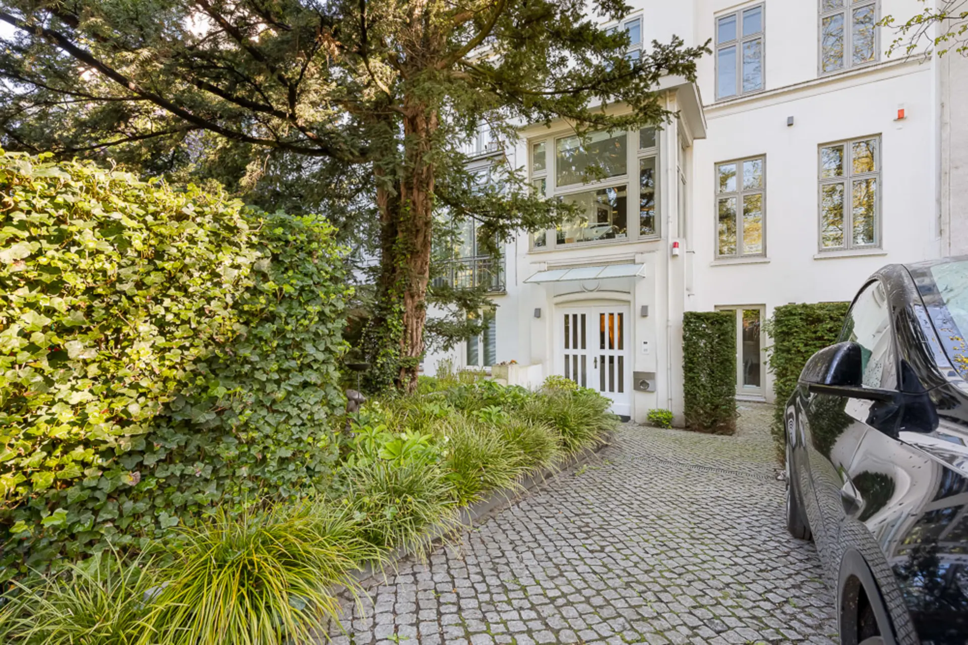 Eingang eines weißen Wohnhauses mit Kopfsteinpflasterweg, Auto und viel Grün vorne.