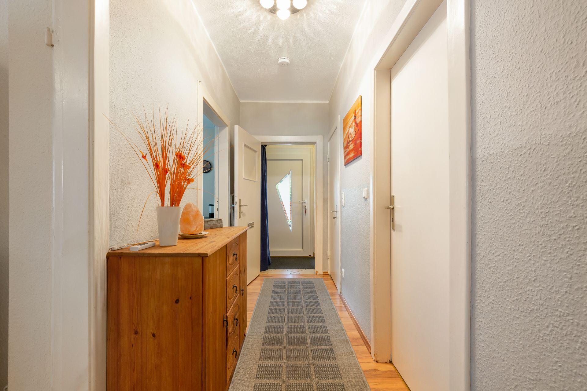 Heller Flur mit Holzkommode, Teppichläufer und Dekoration, mehrere Türen an den Seiten.