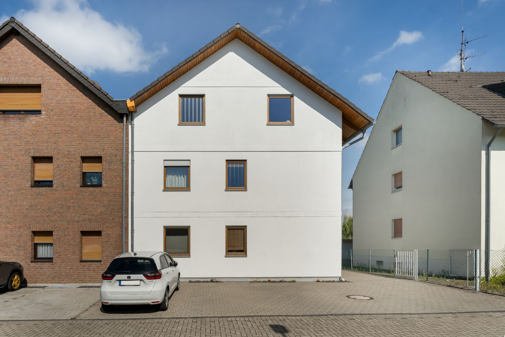 Weiße Wohnhausfassade mit Holzdach, mehreren Fenstern und Parkplatz mit Autos davor.