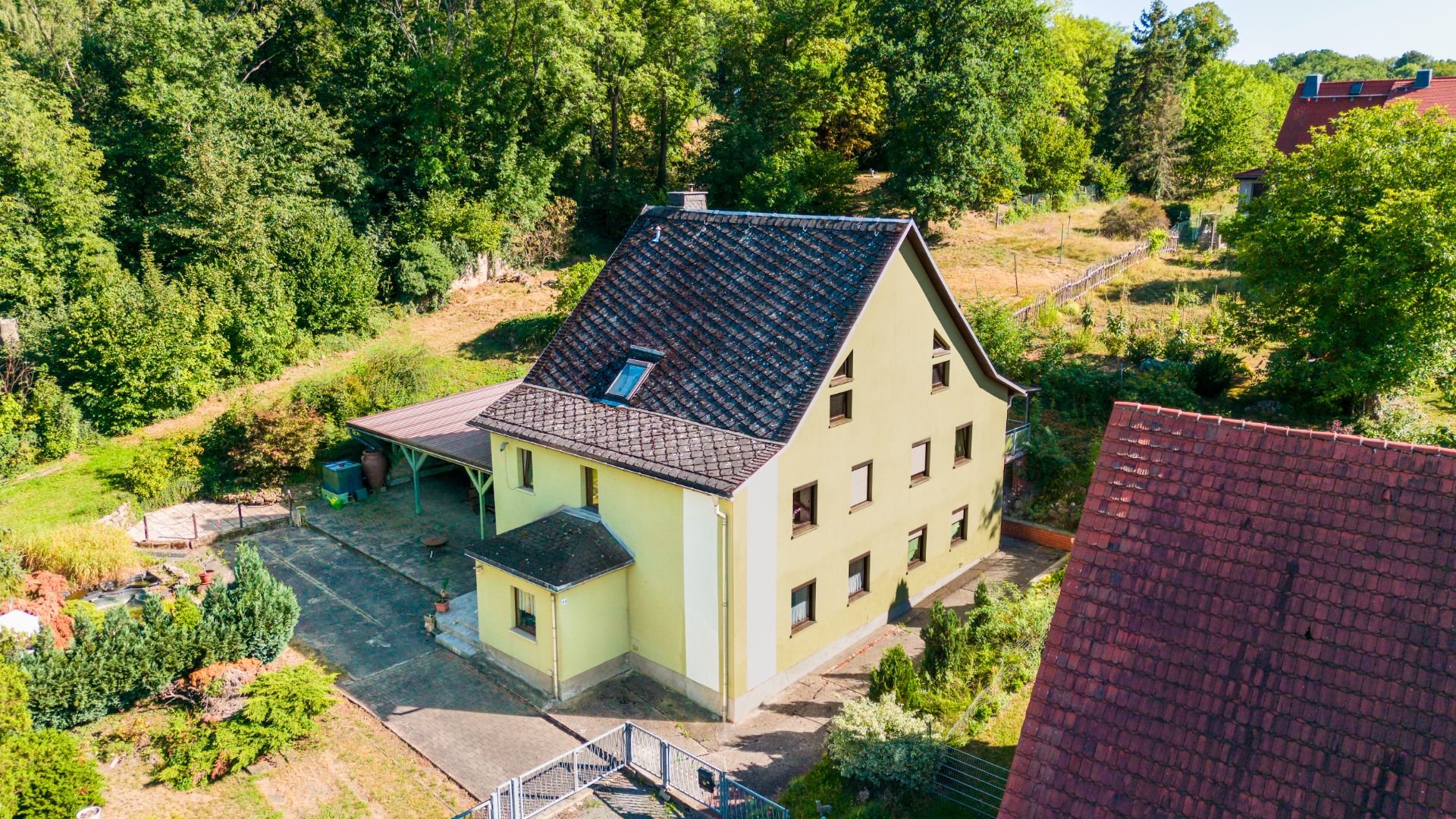 Zweistöckiges gelbes Haus mit dunklem Satteldach, umgeben von Garten und Bäumen.