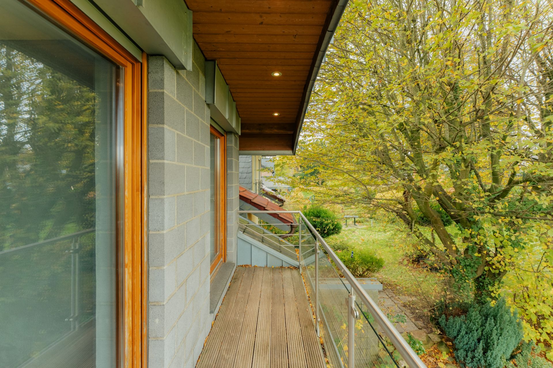 Balkon mit Holzboden, Glasgeländer und Blick auf Bäume und Garten.