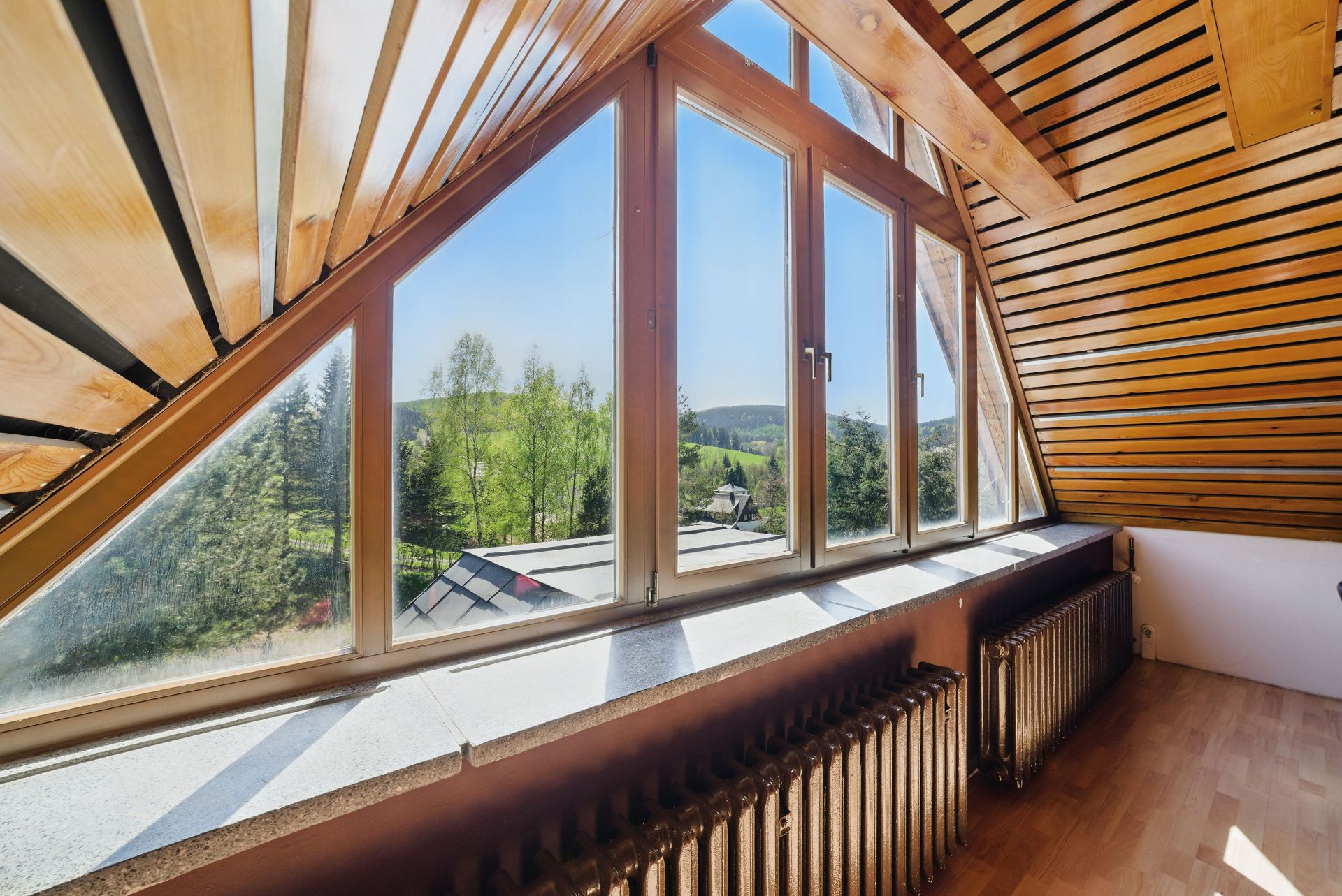 Dachgeschossraum mit großen Fenstern, Holzverkleidung und Blick ins Grüne.