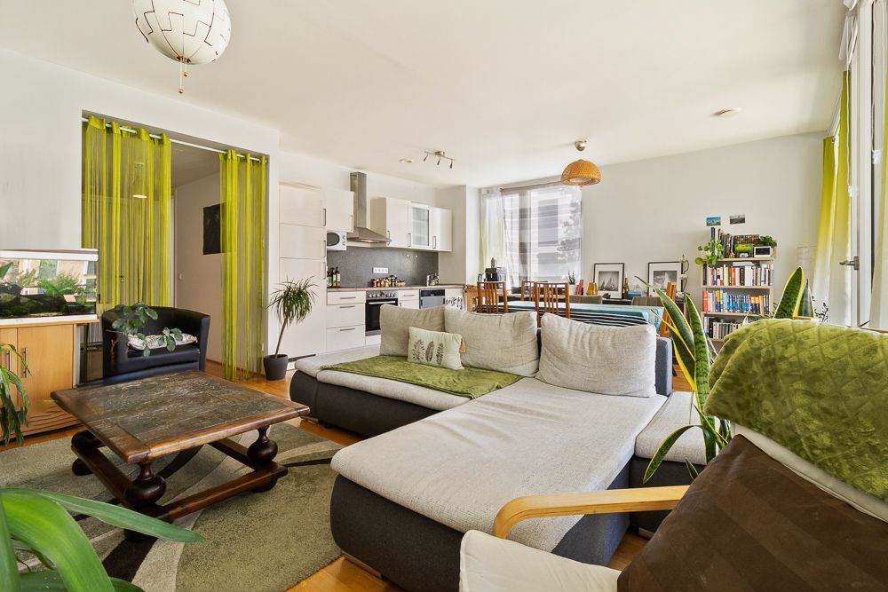 Helles Wohnzimmer mit Sofa, Couchtisch, offenen Küchenbereich und vielen Zimmerpflanzen.